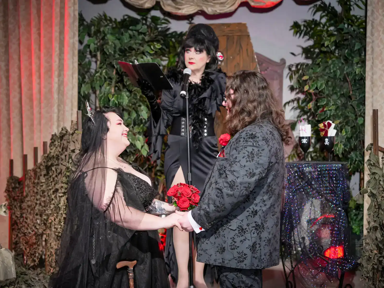 Couple holding hands during gothic wedding ceremony with officiant reading vows on stage.