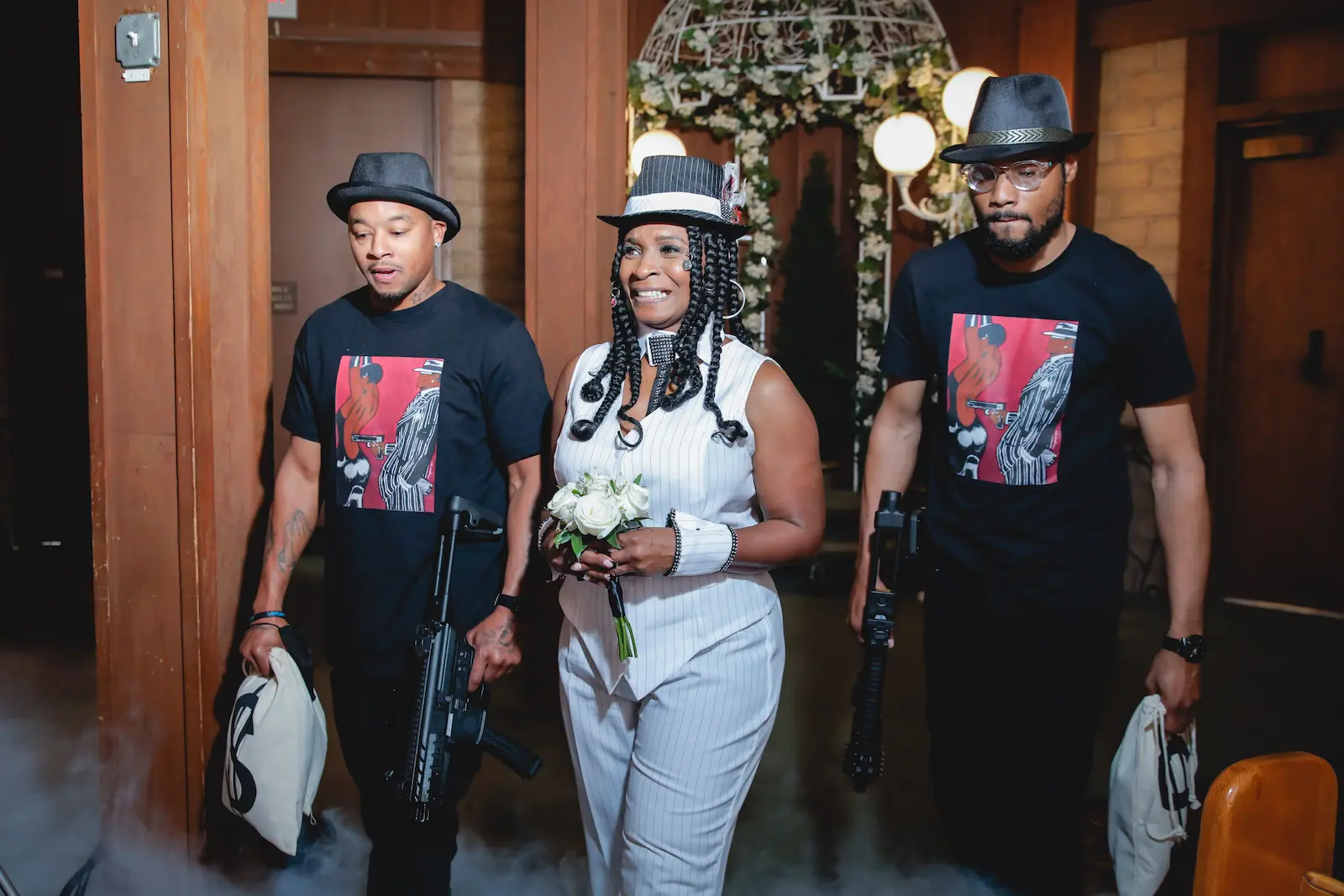 Bride in white pinstripe gangster outfit escorted down aisle by two groomsmen with prop guns at Las Vegas chapel