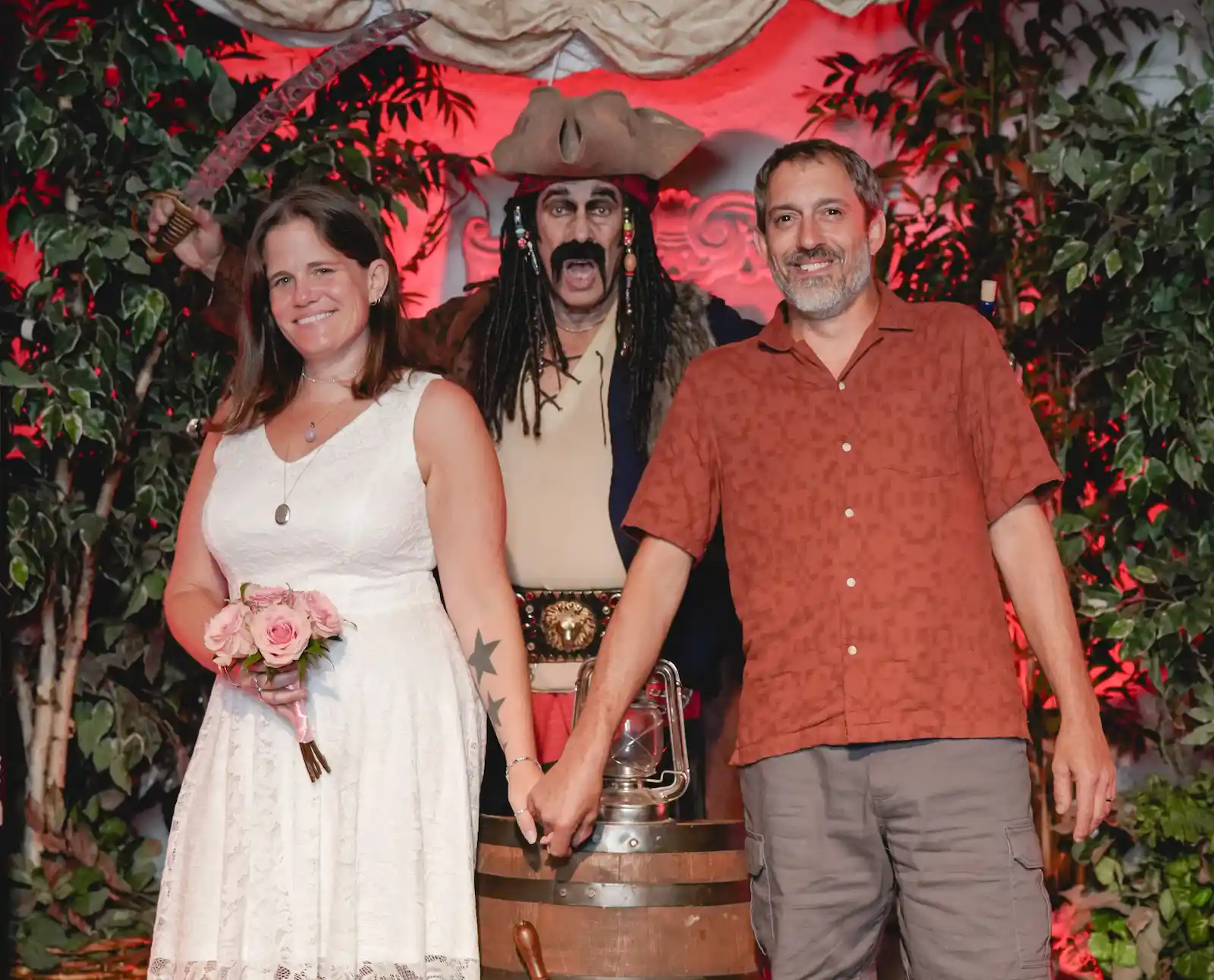 Bride and groom posing with a pirate character behind a barrel ship wheel at a Las Vegas pirate wedding venue.