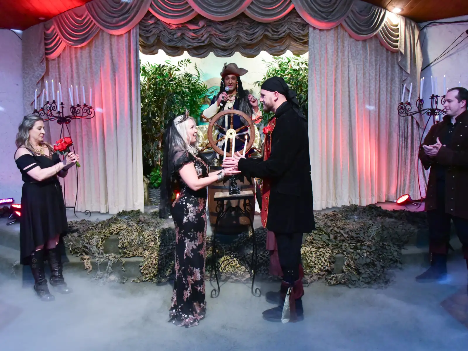 A bride and groom in steampunk wedding attire posing with a pirate-themed officiant and a decorative skeleton at a fantasy wedding venue.