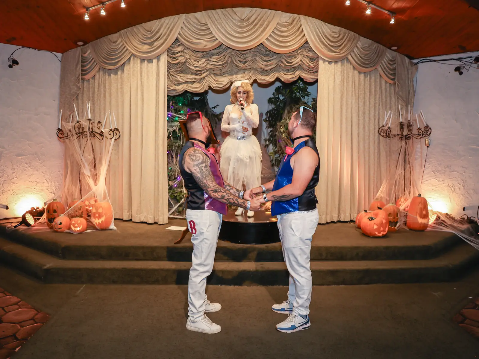 same-sex couple holding hands during halloween themed wedding ceremony with pumpkins and performer officiant