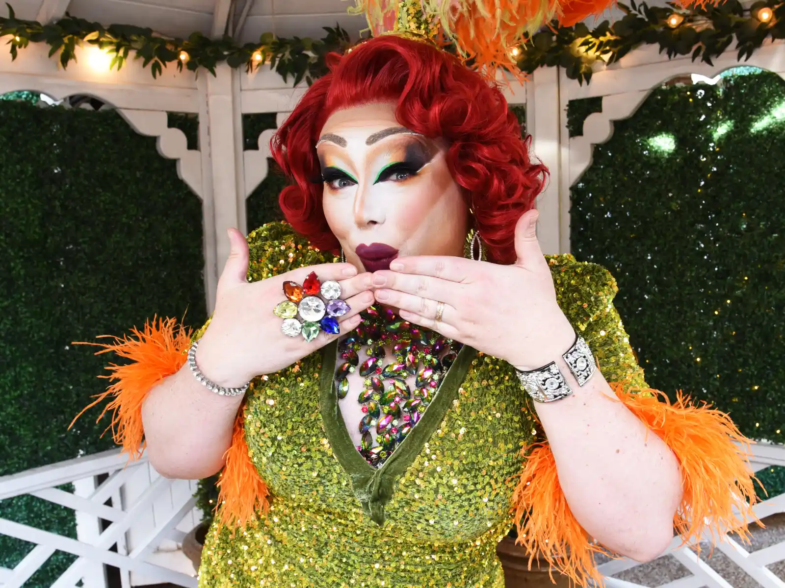 A drag performer with red hair and a green sequin dress blowing a kiss, featuring orange feather trim and a large colorful flower ring.
