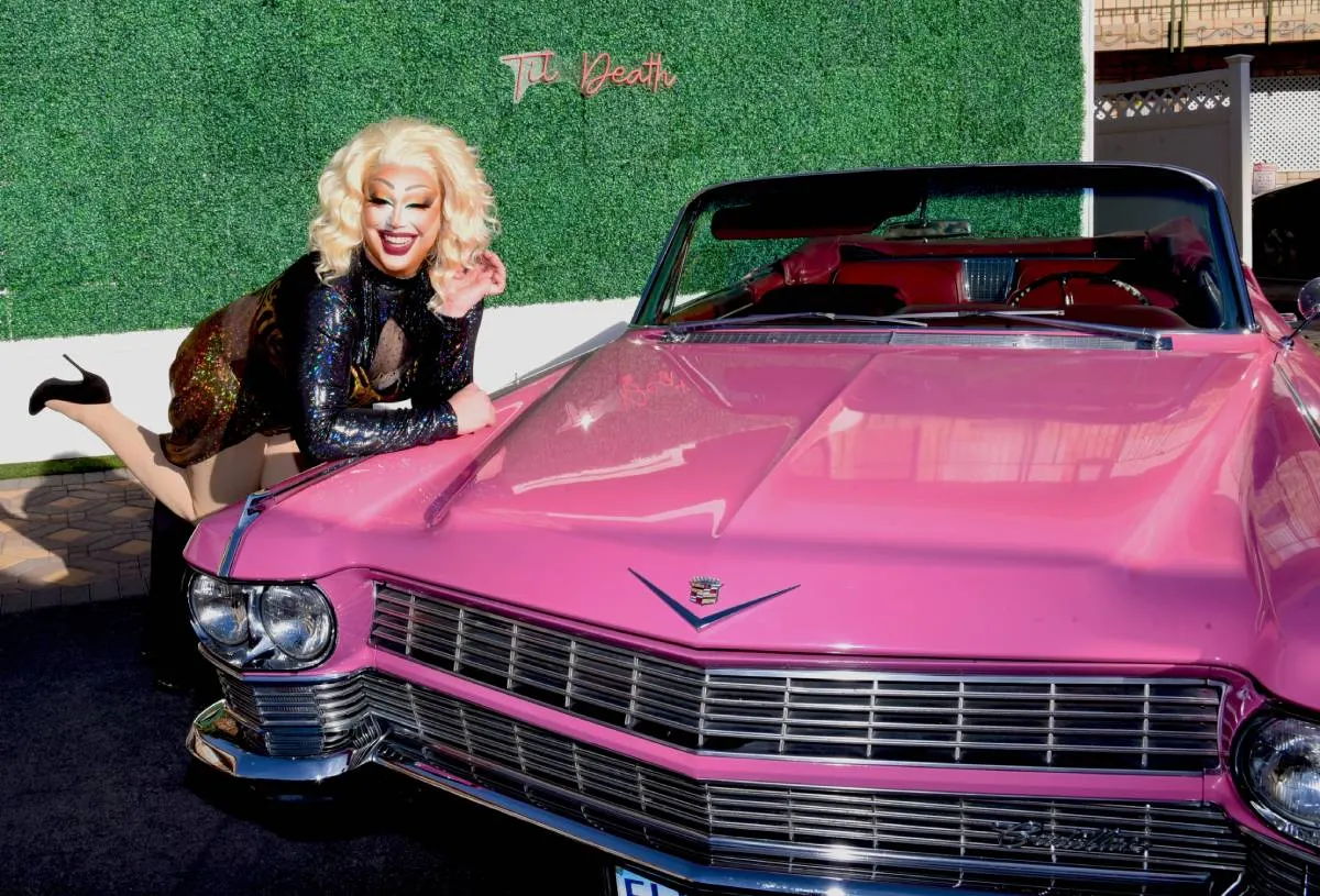 A drag queen with blonde hair and a black sequin dress posing next to a vintage pink Cadillac convertible in front of a green hedge with a "Til Death" sign.