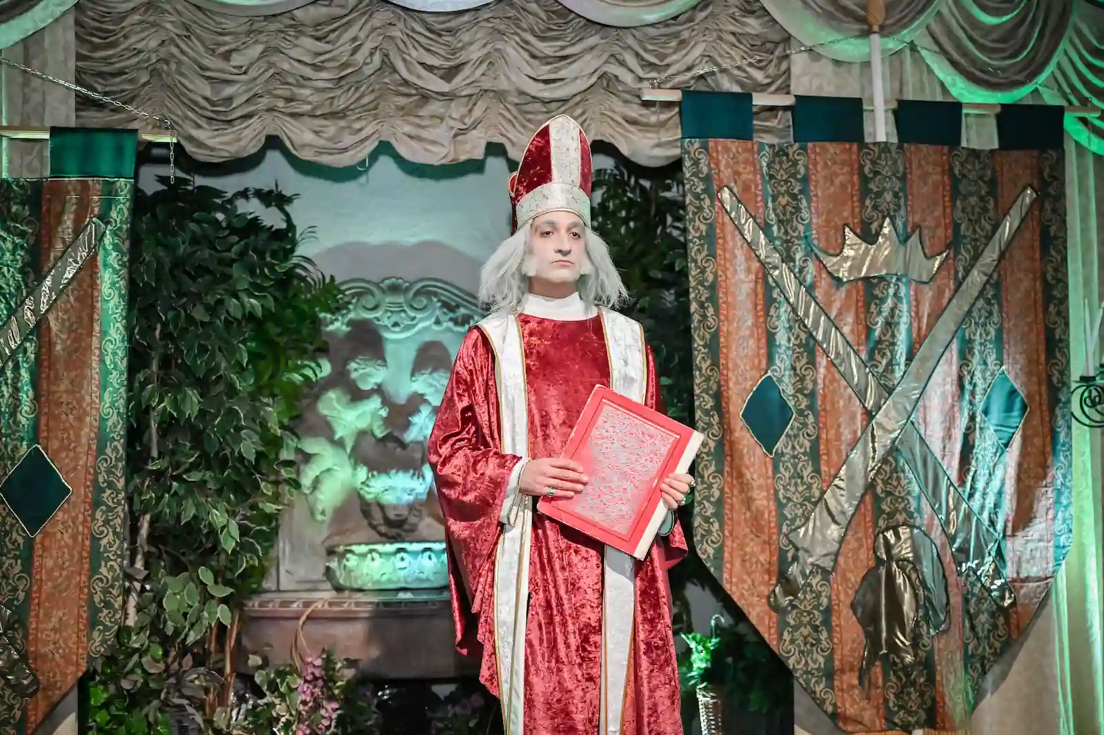 Medieval bishop officiant in a red velvet robe holding a ceremonial book before royal banners at a Las Vegas wedding chapel.