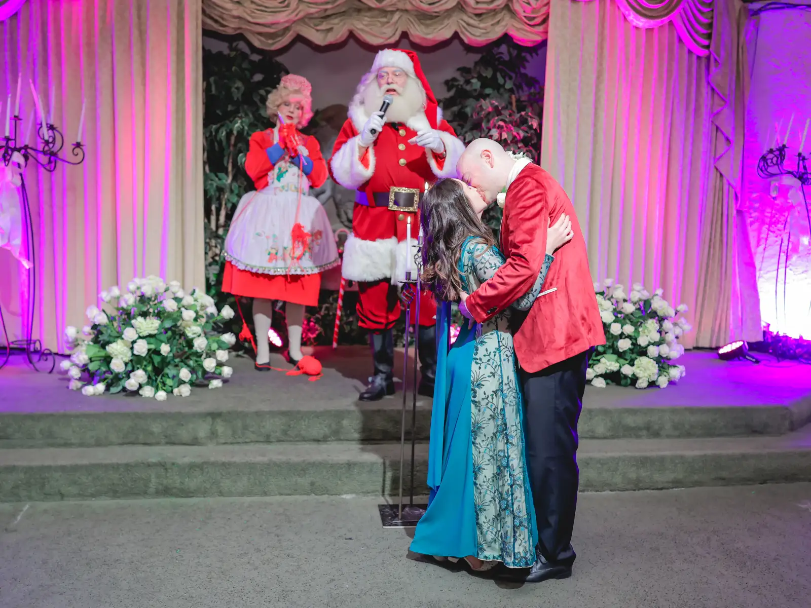 A couple kisses on a stage with vibrant pink lighting, with Santa Claus holding a microphone and Mrs. Claus standing in the background.