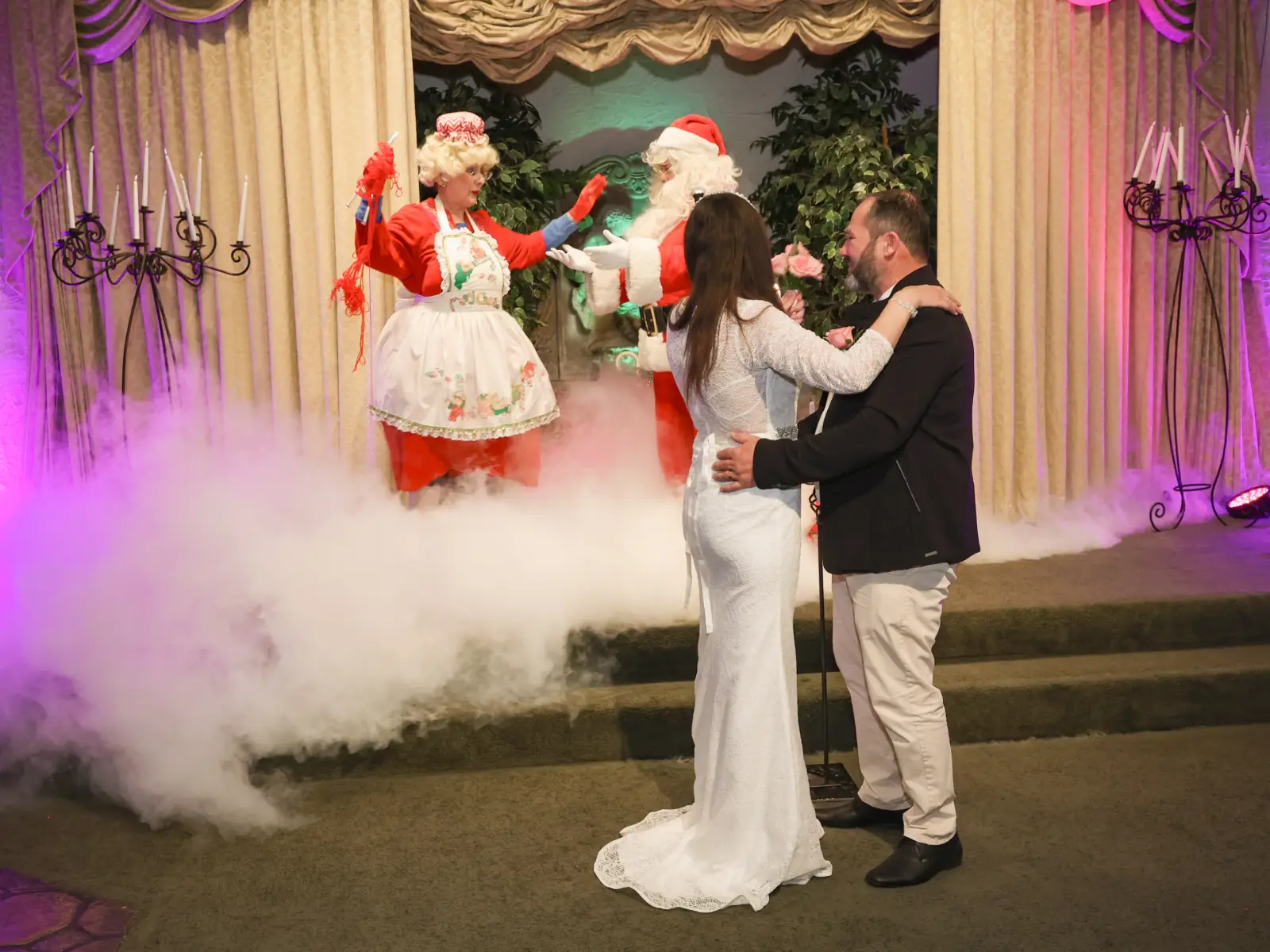 A bride and groom in a warm embrace dance on a smoke-filled stage, with Santa and Mrs. Claus performing in the background.
