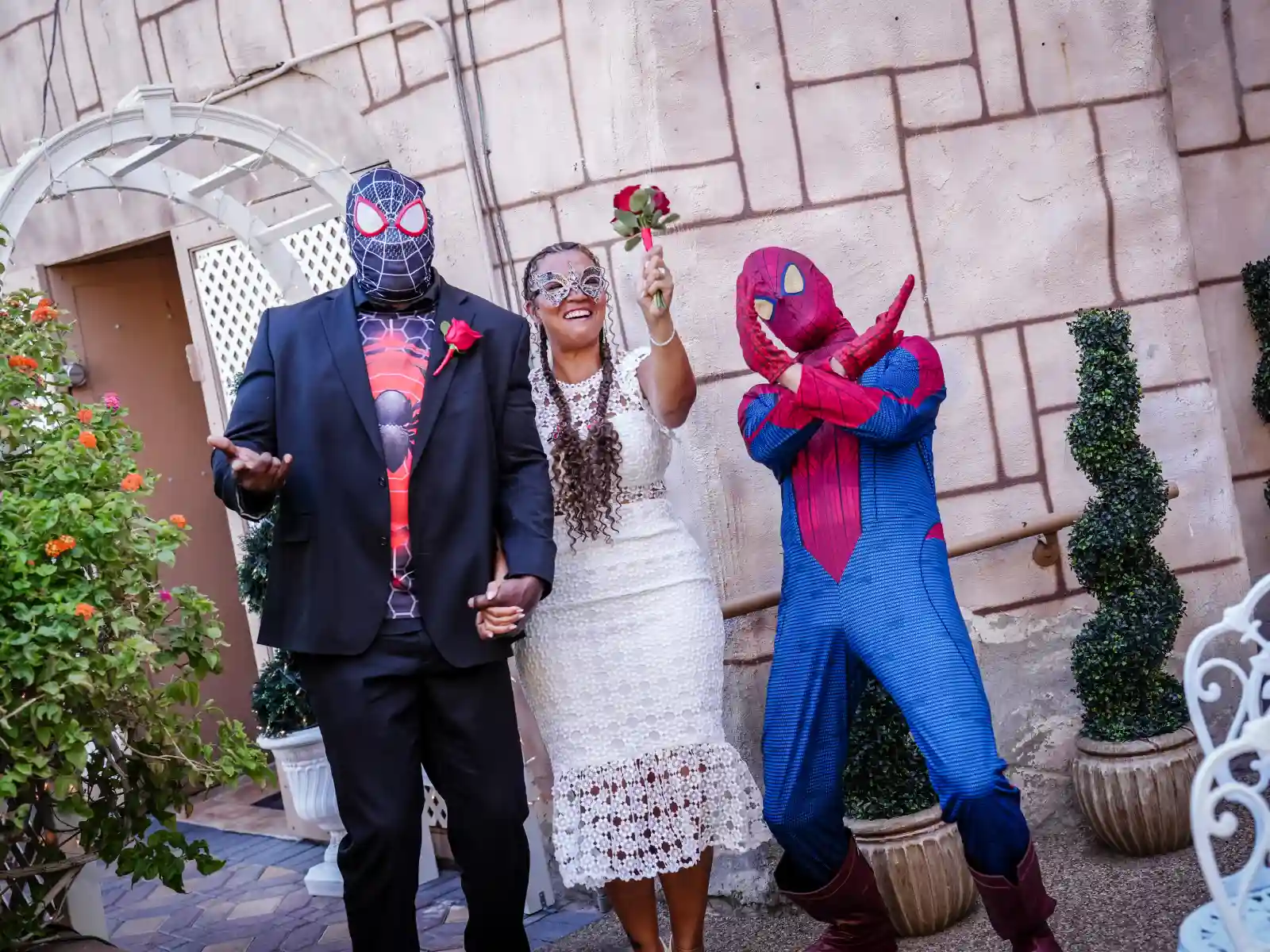 A groom wearing a Miles Morales Spider-Man mask and suit under a black blazer poses with his bride, who wears a white lace dress and silver mask, alongside a classic Spider-Man performer.