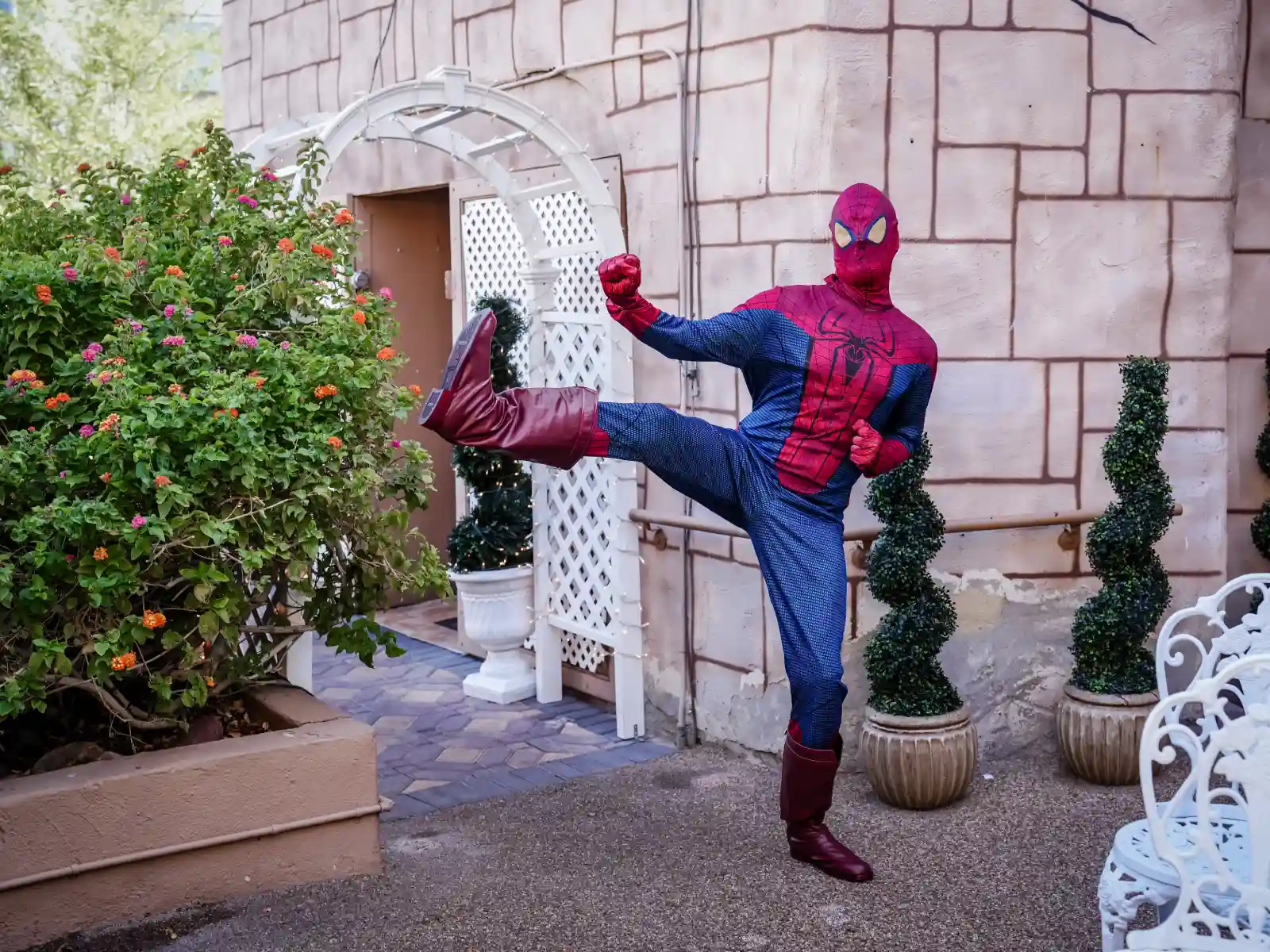 A Spider-Man impersonator performing a high side kick outside a wedding venue. The character is wearing red boots and a detailed spandex suit against a stone wall background.