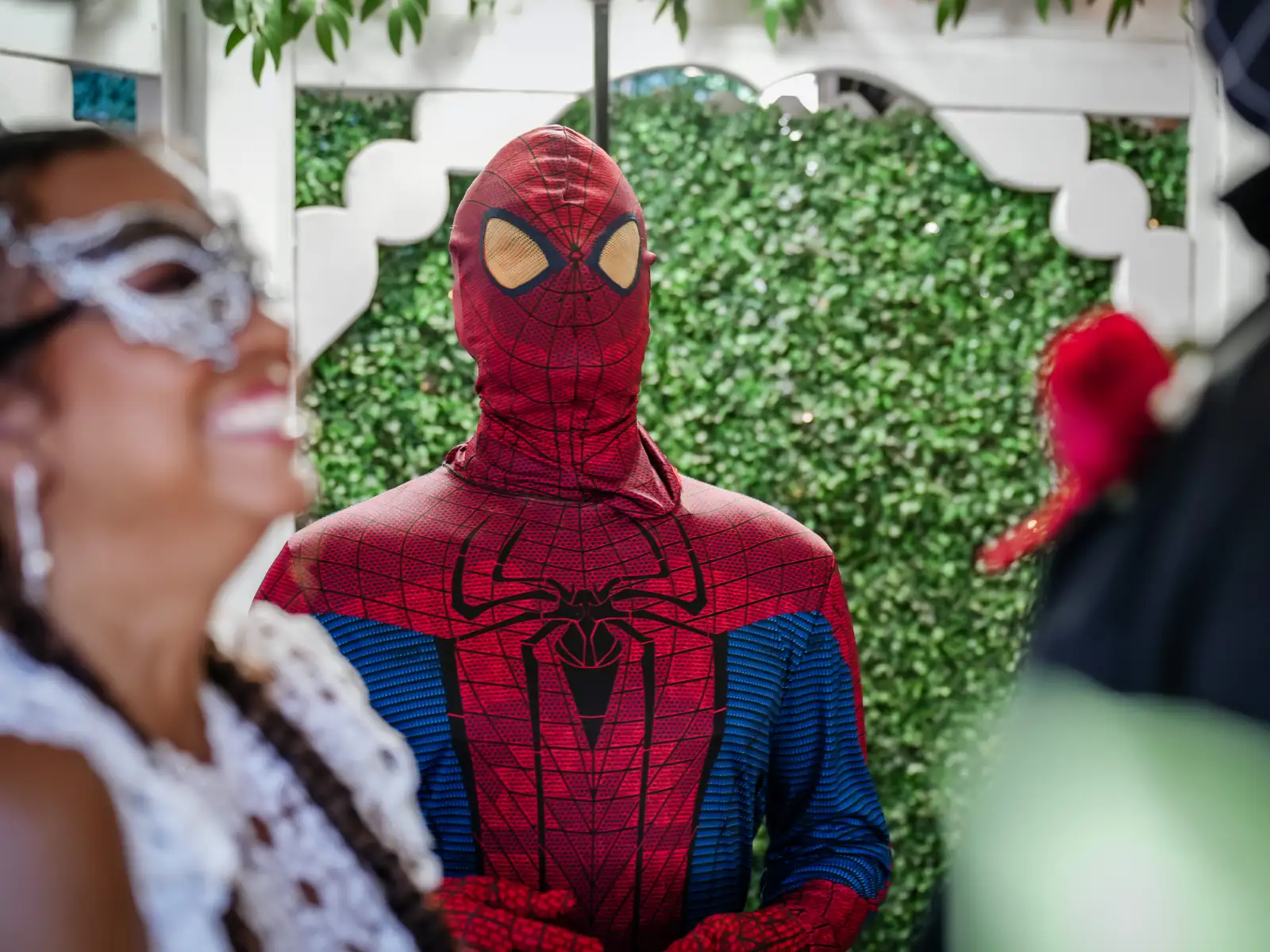 A close-up portrait of a Spider-Man impersonator in a classic red and blue suit standing behind a smiling wedding guest wearing a decorative silver masquerade mask.