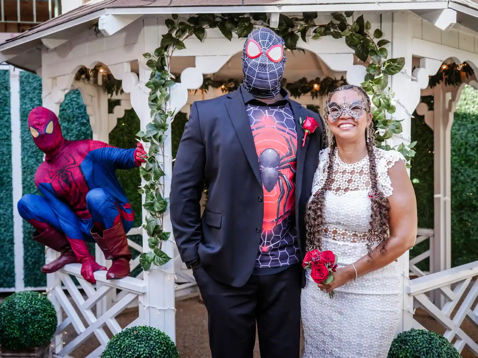 A wedding couple in superhero-themed attire poses in front of a white garden gazebo. The groom wears a Miles Morales mask and the bride a silver eye mask, while a Spider-Man impersonator crouches on the railing in a signature pose.