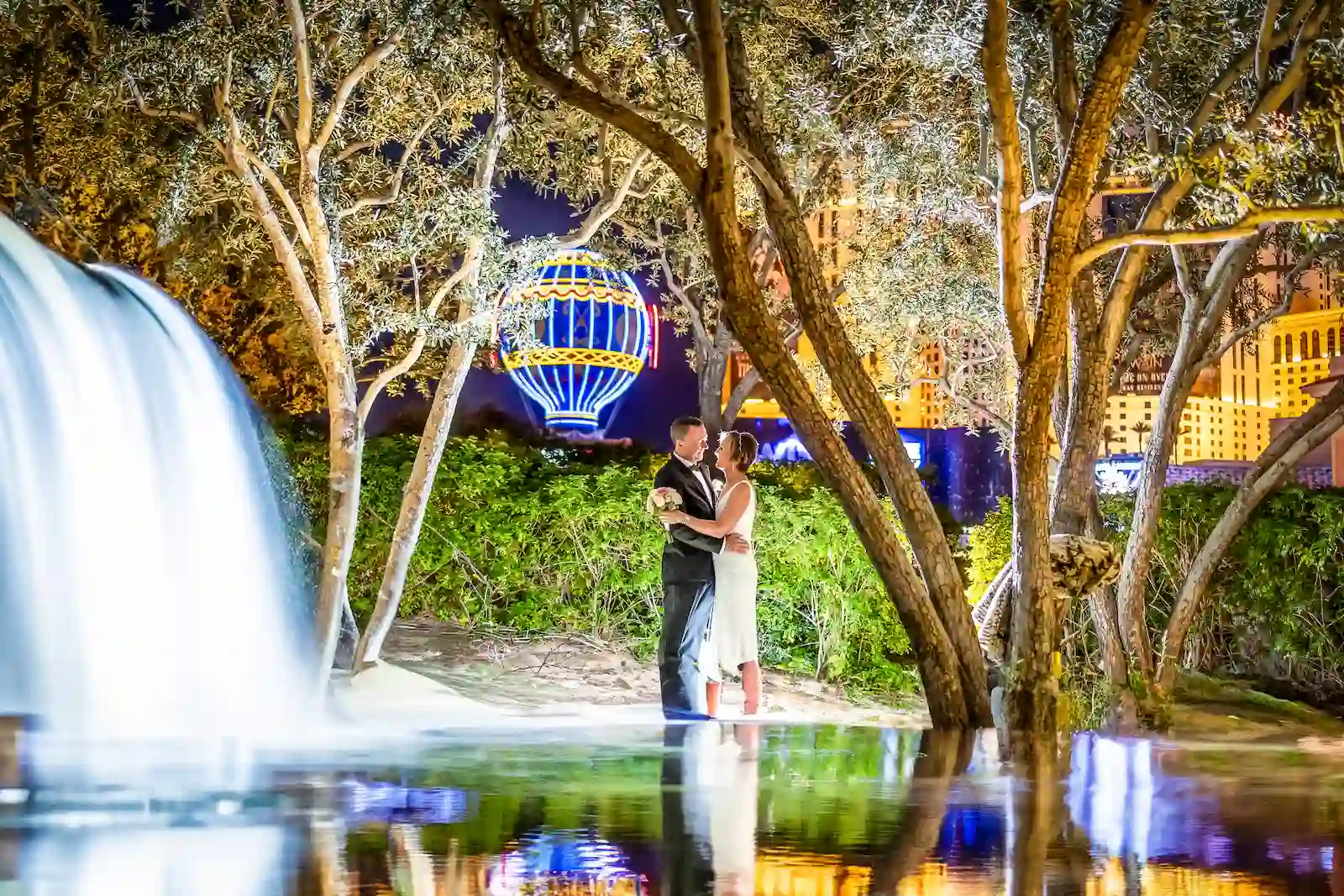 Bride and groom embracing near a waterfall on the Las Vegas Strip at night with the Paris Las Vegas balloon in the background.