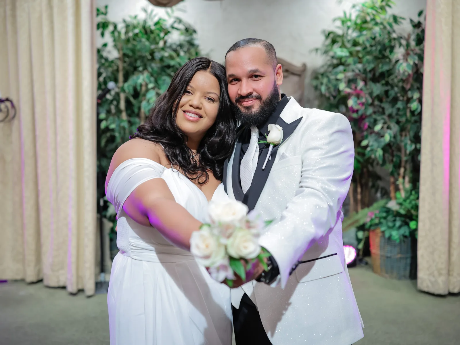 A bride in a sleek, vintage-style beaded gown and a groom in a navy blue suit and brown shoes sharing a romantic moment on a stage. They are framed by heavy beige theater drapes and greenery, with warm stage lighting and wrought-iron candelabras on either side.