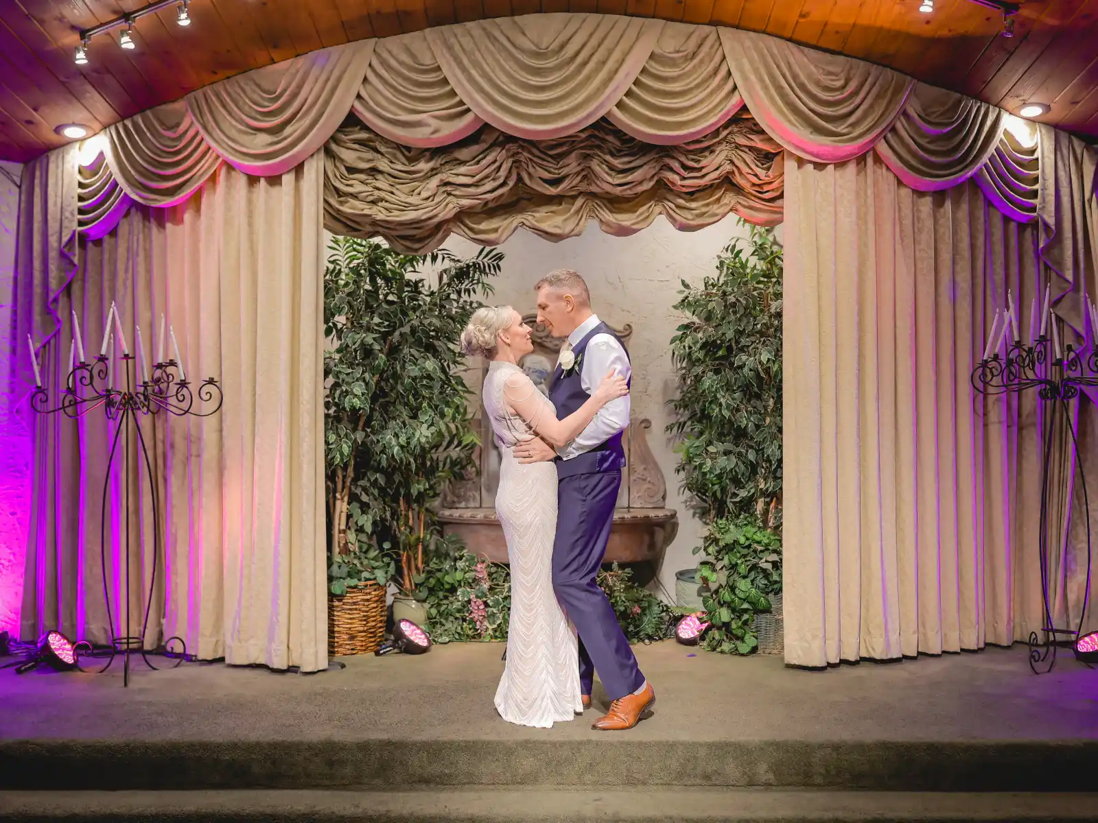 A bride in a sleek, vintage-style beaded gown and a groom in a navy blue suit and brown shoes sharing a romantic moment on a stage. They are framed by heavy beige theater drapes and greenery, with warm stage lighting and wrought-iron candelabras on either side.