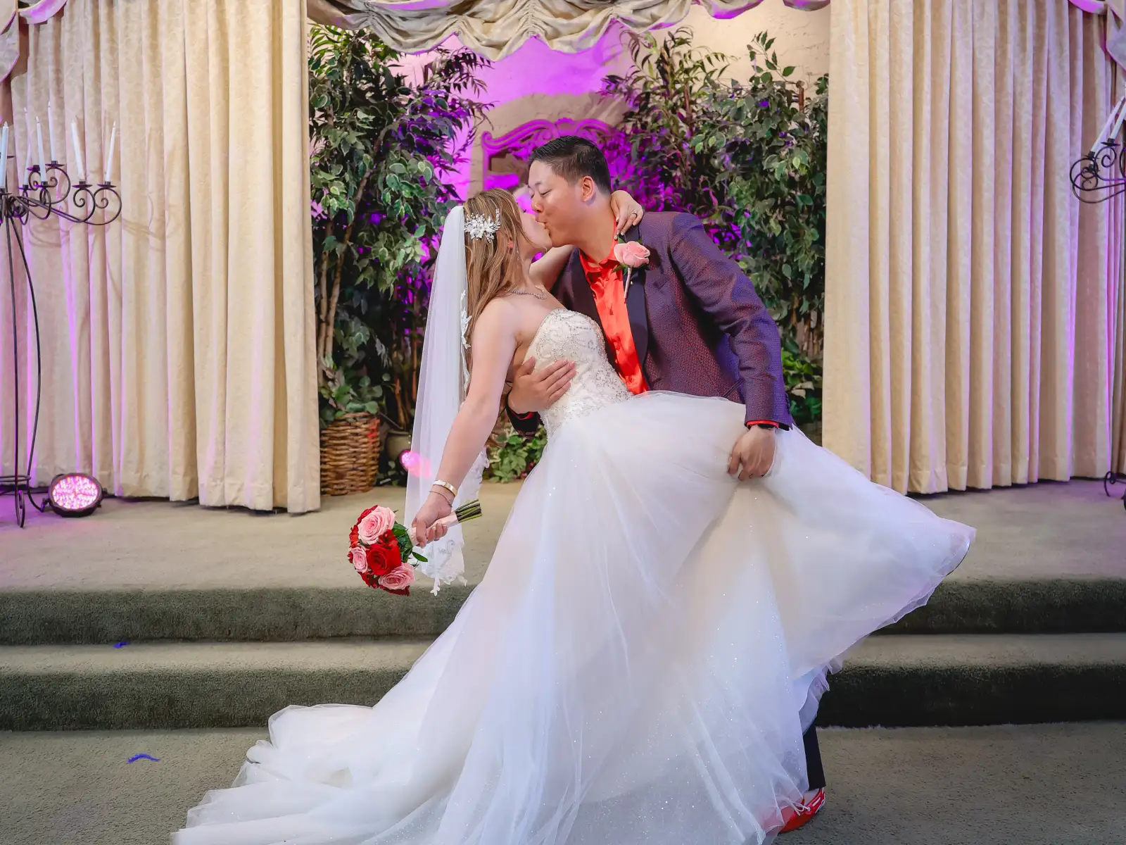 A groom in a stylish purple suit dips and kisses his bride in a wedding chapel, featuring purple ambient lighting and lush green backdrop plants.