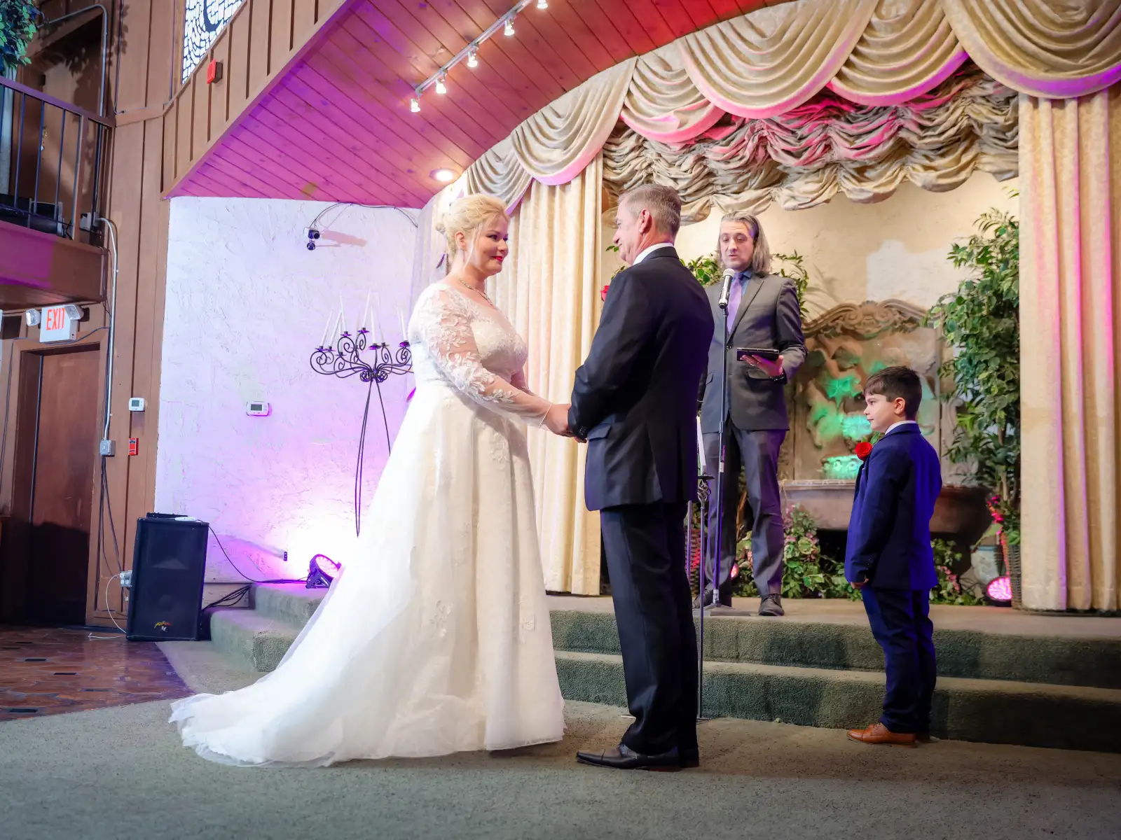 A bride in a long-sleeved lace gown and a groom in a black tuxedo holding hands during their wedding ceremony on a stage. An officiant stands behind a microphone, and a young ring bearer in a navy suit looks on from the side.