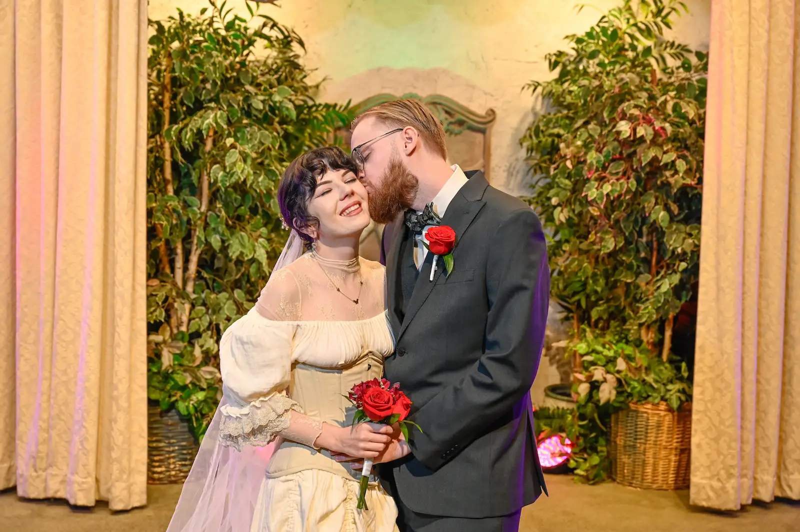 Groom kissing bride on the cheek at Viva Las Vegas Wedding Chapel, bride holding red rose bouquet.