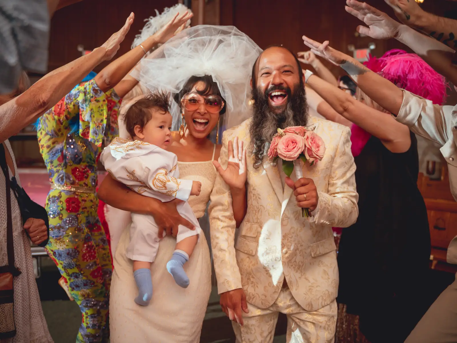 A vibrant and eclectic wedding photo of a bride in oversized sunglasses holding a baby, alongside a groom with a long beard in a gold patterned suit, laughing as guests reach out toward them.