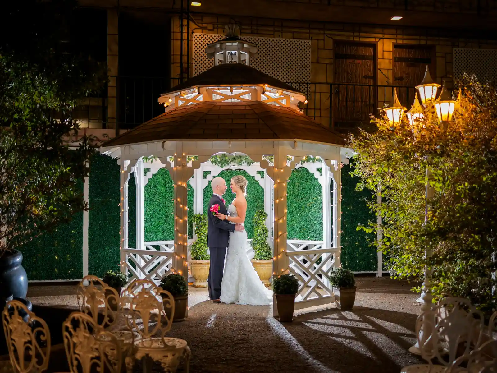 A romantic night shot of a bride and groom gazing at each other inside a white garden gazebo decorated with warm fairy lights and surrounded by greenery.