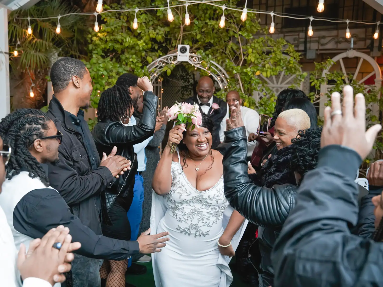 A joyful bride in an embellished white gown raising her pink rose bouquet as she walks through a cheering crowd of wedding guests under string lights.