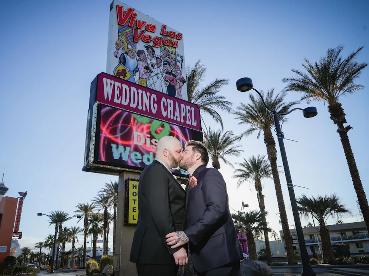 Grooms kissing beneath Viva Las Vegas Wedding Chapel sign with palm trees and blue sky.