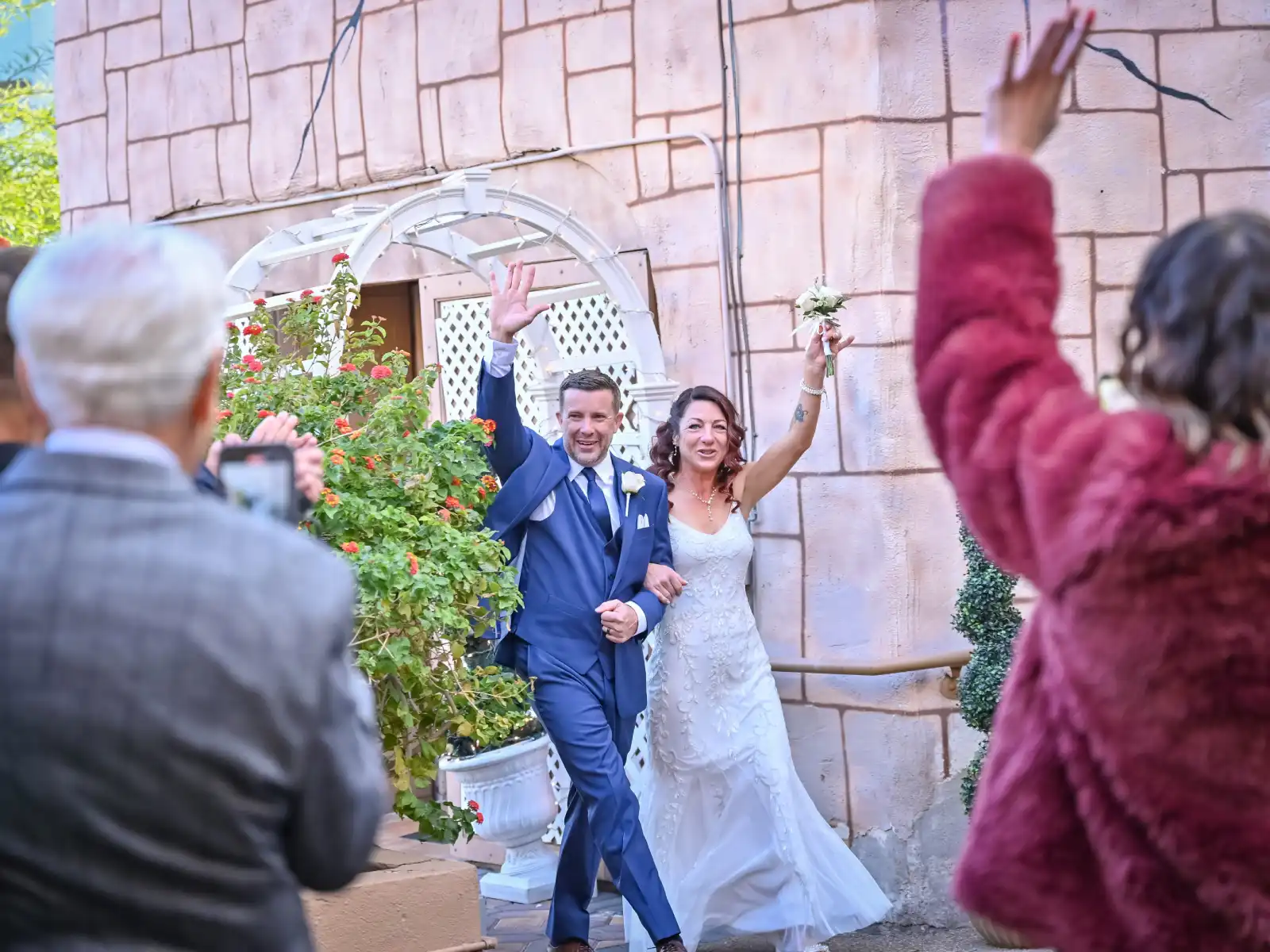 A happy bride and groom holding hands and laughing during their outdoor wedding ceremony. The bride is wearing a sleeveless lace gown, and the groom is in a dark suit with a white rose boutonniere. They are standing in front of a white garden gazebo decorated with greenery and pink flowers, with guests seated in the background.