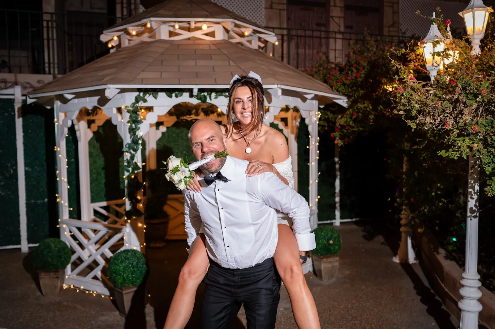 Bride riding piggyback on groom with rose at romantic Las Vegas gazebo wedding at night