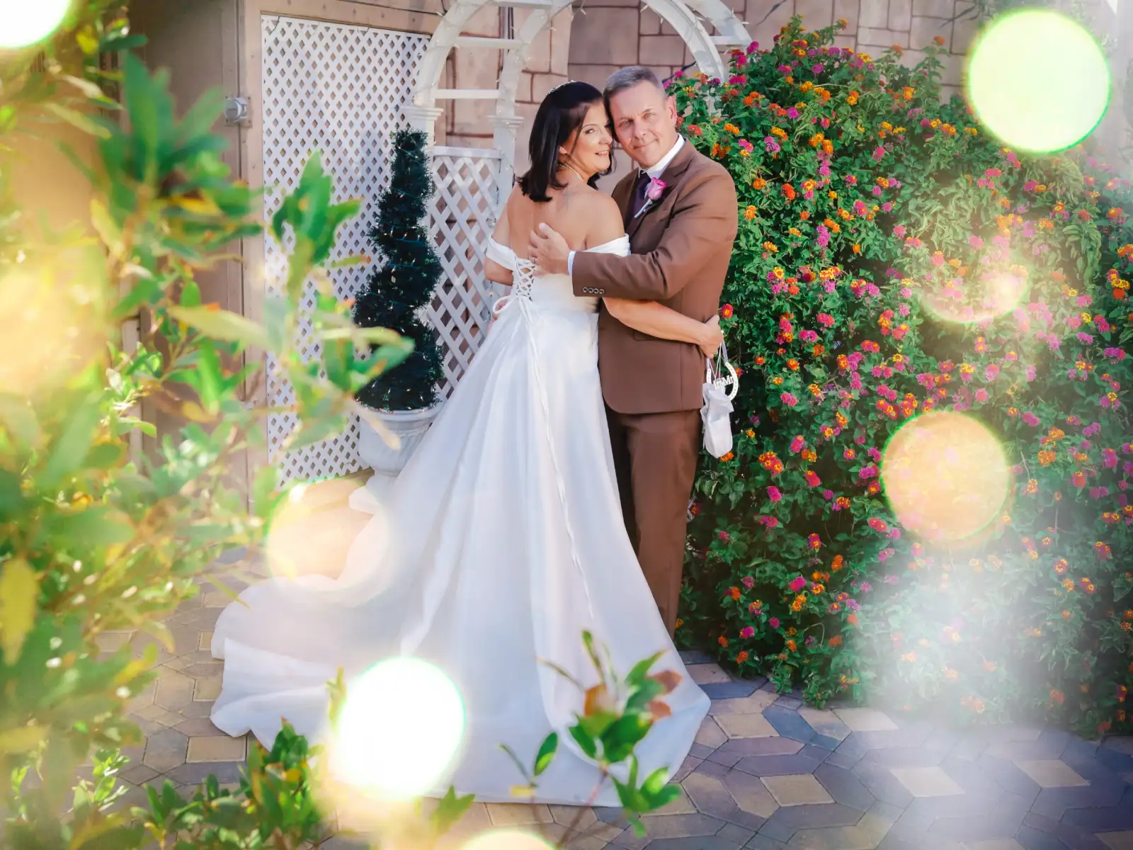 A happy bride in a white lace dress with a long train and a groom in a navy blue suit and brown shoes embracing inside a white garden gazebo, gazing into each other
