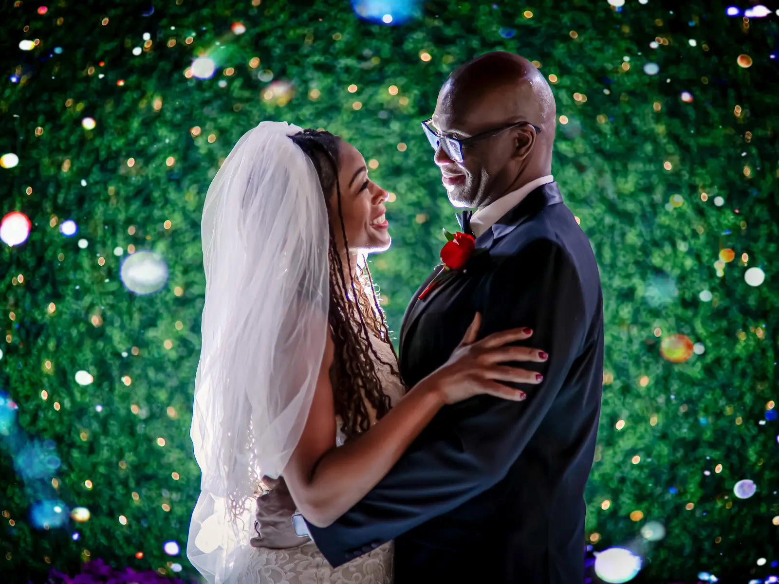 A close-up romantic portrait of a bride with long braids and a groom with glasses and a red rose boutonniere, looking into each other