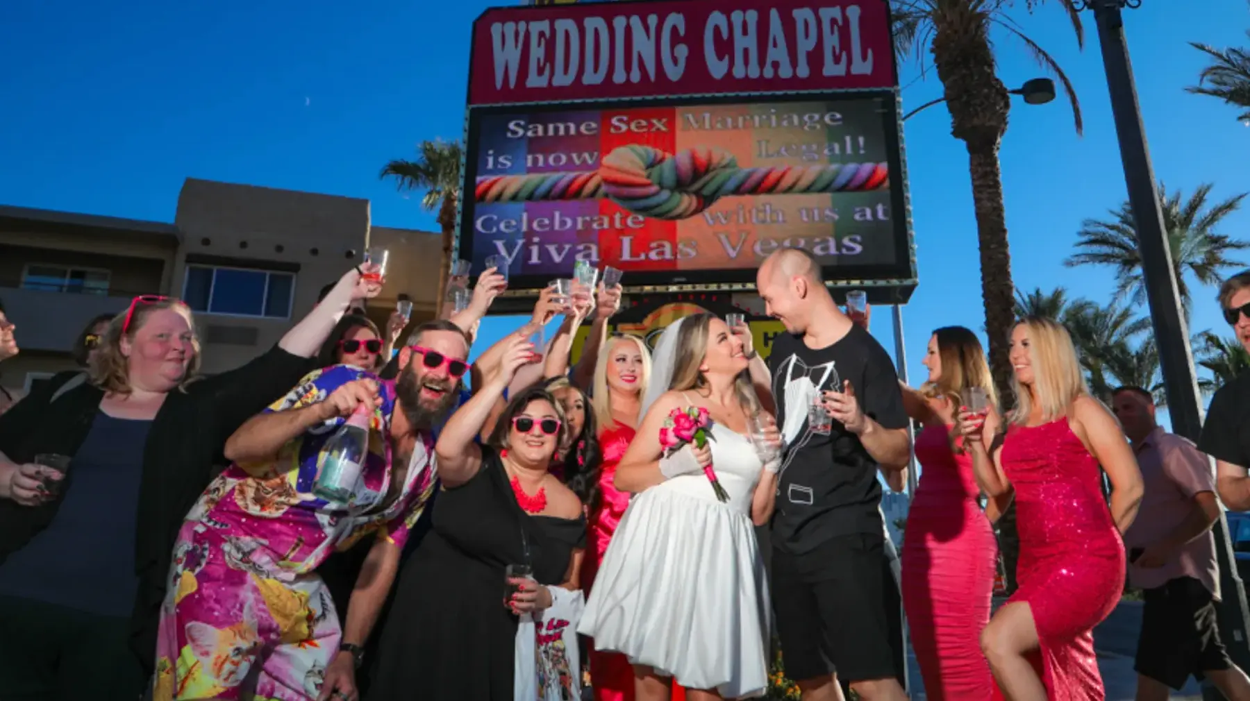 A celebratory outdoor wedding scene at the Viva Las Vegas Wedding Chapel; guests in colorful attire raise glasses in a toast while a bride in a short dress and a groom in a tuxedo-print t-shirt smile under a marquee.
