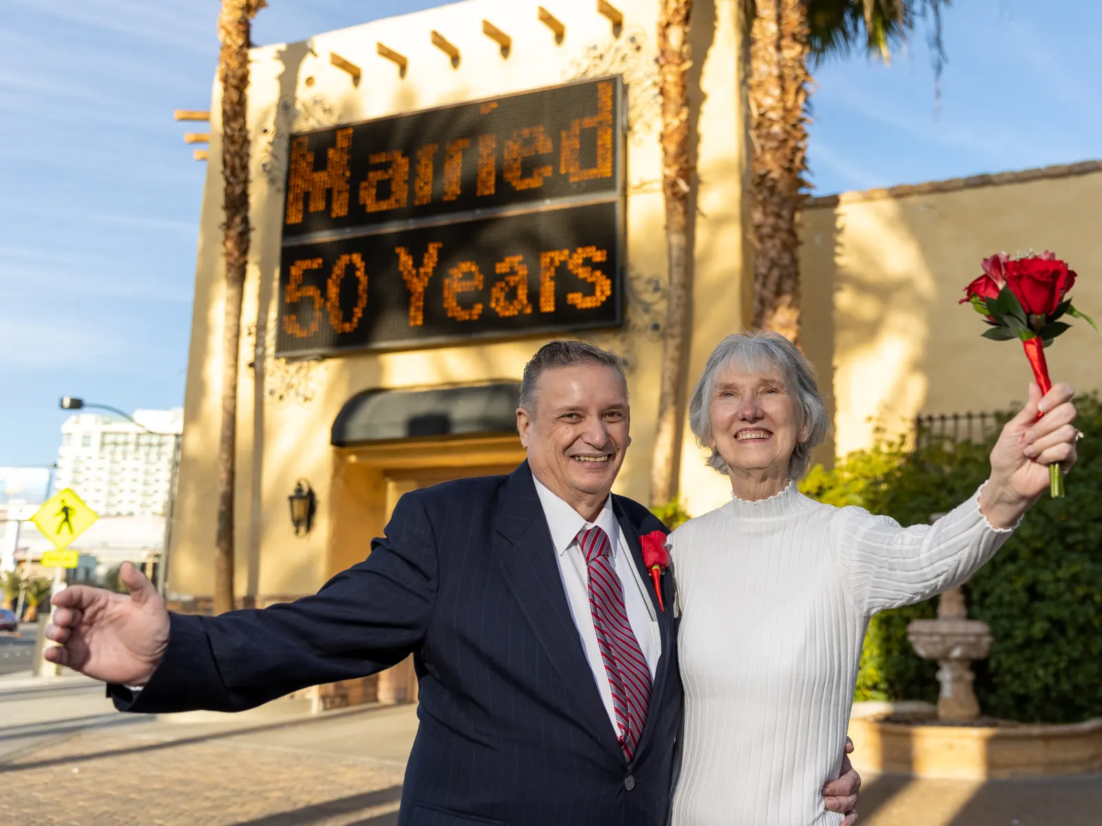 A happy senior couple smiling and celebrating in front of a wedding chapel marquee that reads "Married 50 Years," with the woman holding up a small bouquet of red roses.