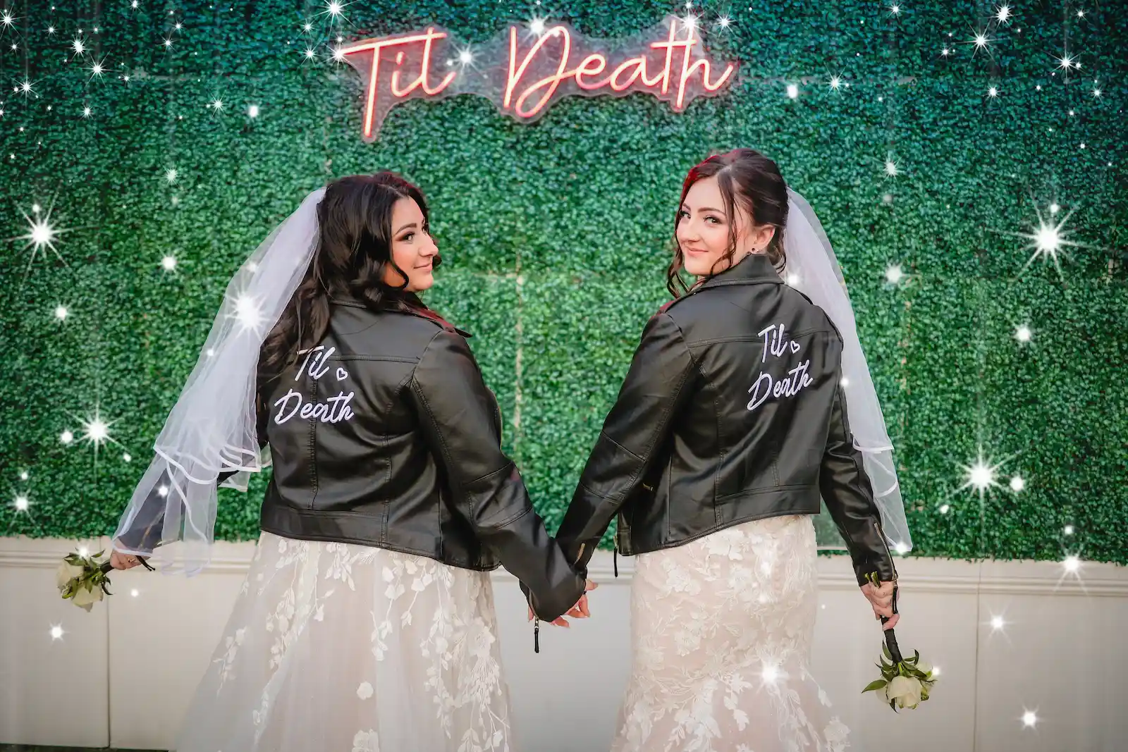 Two brides in matching "Til Death" leather jackets and veils holding hands in front of a hedge wall at Viva Las Vegas Wedding Chapel.