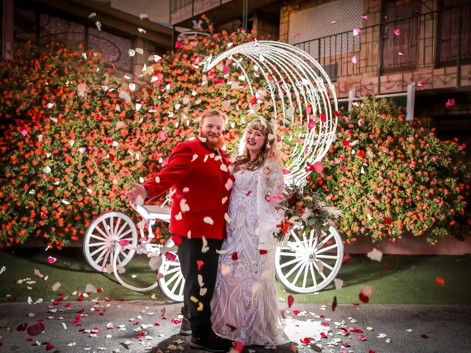 A joyful groom in a red velvet suit and a bride in a lace gown standing under a shower of falling rose petals with a decorative white carriage and floral backdrop.