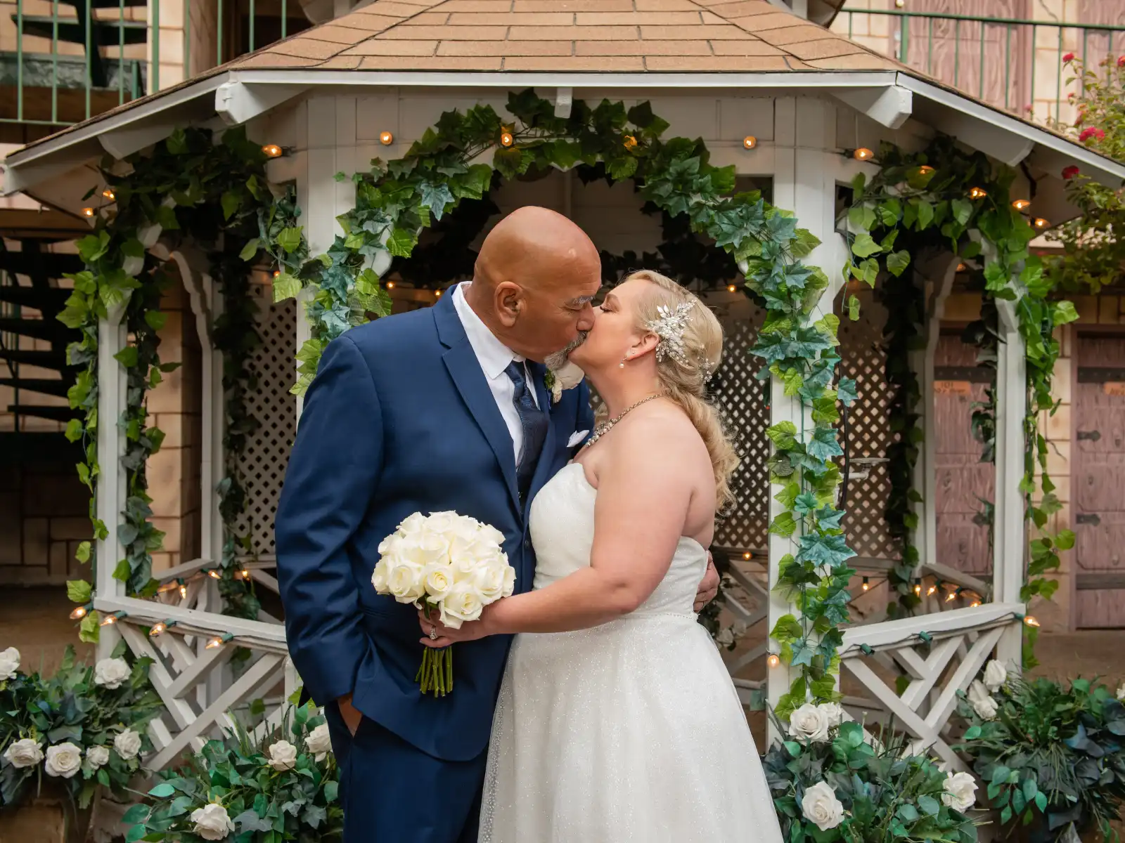 An intimate shot of a bride holding a white rose bouquet kissing her groom inside a white garden gazebo decorated with ivy and fairy lights.