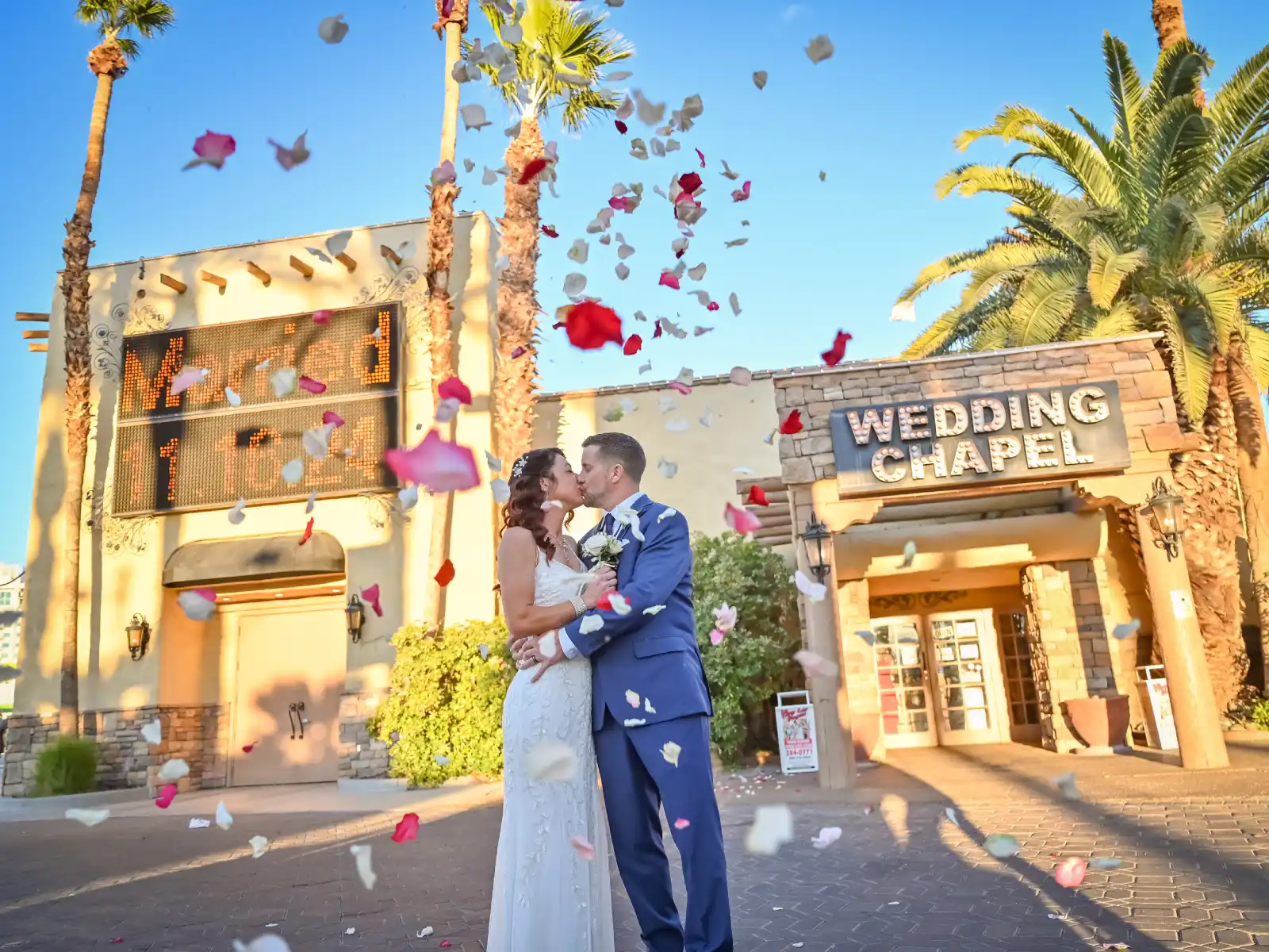 A bride and groom kissing in front of a Las Vegas wedding chapel with rose petals falling in the air and palm trees under a clear blue sky.
