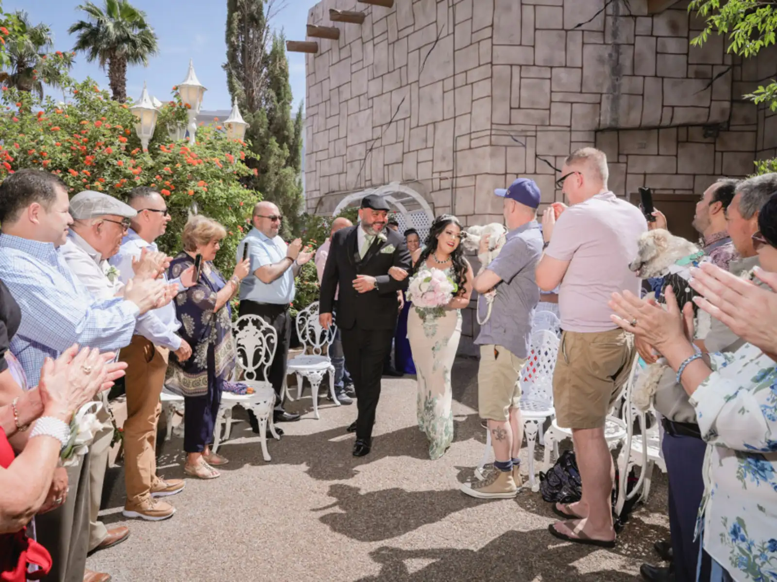 A newly married couple walking down the aisle at an outdoor wedding ceremony while guests clap and cheer in a sunlit courtyard