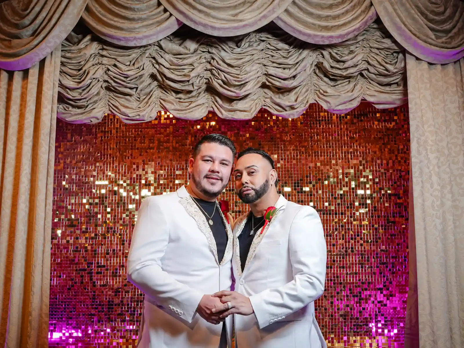 Two grooms in white suits hold hands in front of a sequin wall.