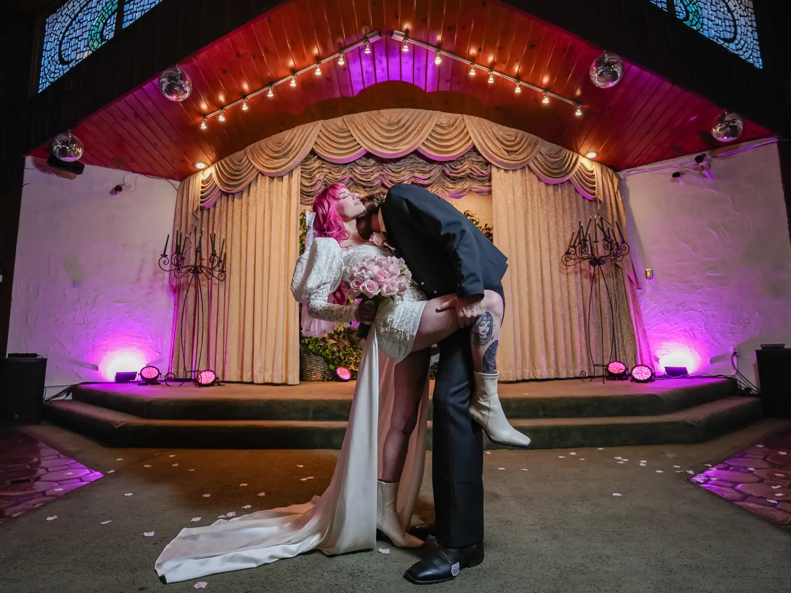 A couple performs a dramatic dip kiss on a stage with disco balls and purple lighting.