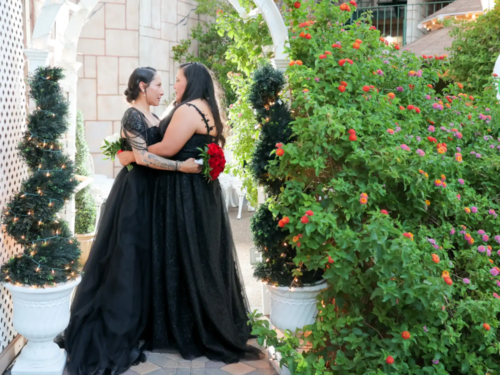 Two brides wearing elegant black wedding gowns embrace in an outdoor garden setting, framed by white arches and spiral topiary trees with twinkle lights.