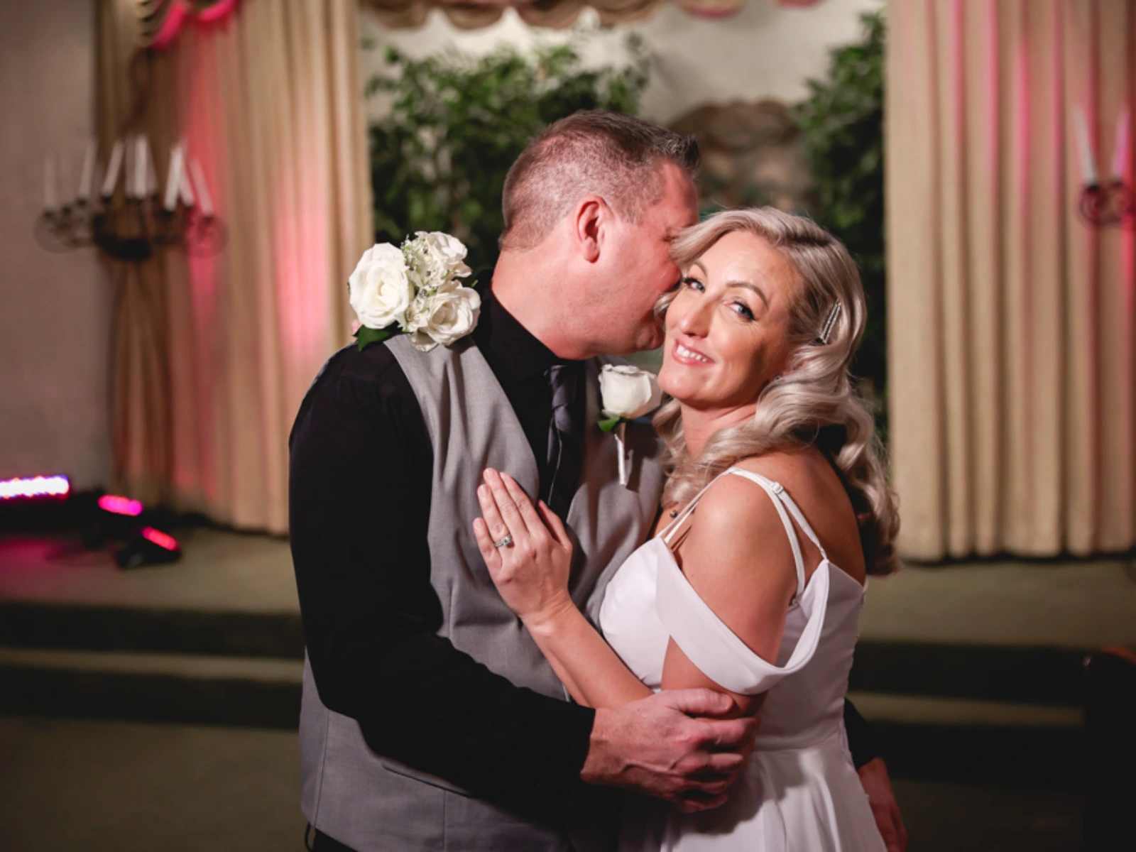 A bride in a white gown smiles at the camera while her groom in a grey vest whispers in her ear during their chapel wedding ceremony.