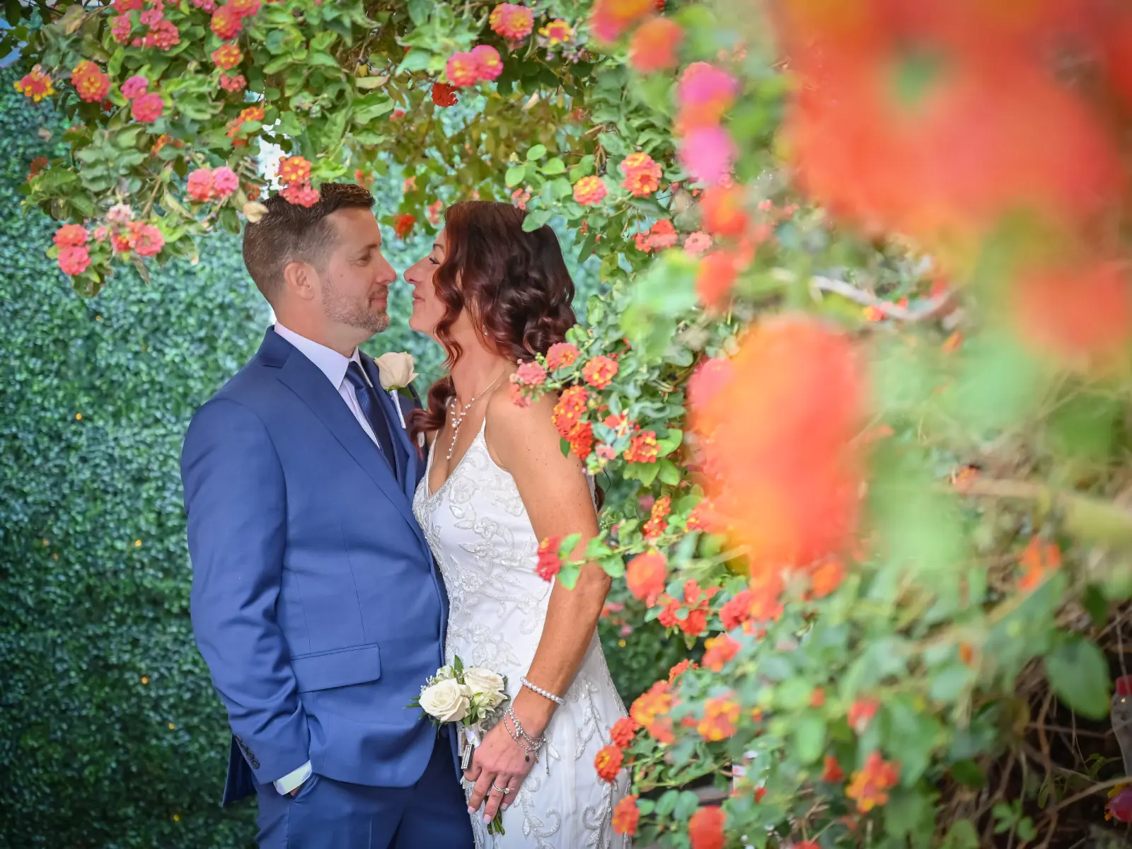 A romantic shot of a bride and groom about to kiss, beautifully framed by vibrant orange and pink flowers in an outdoor garden.
