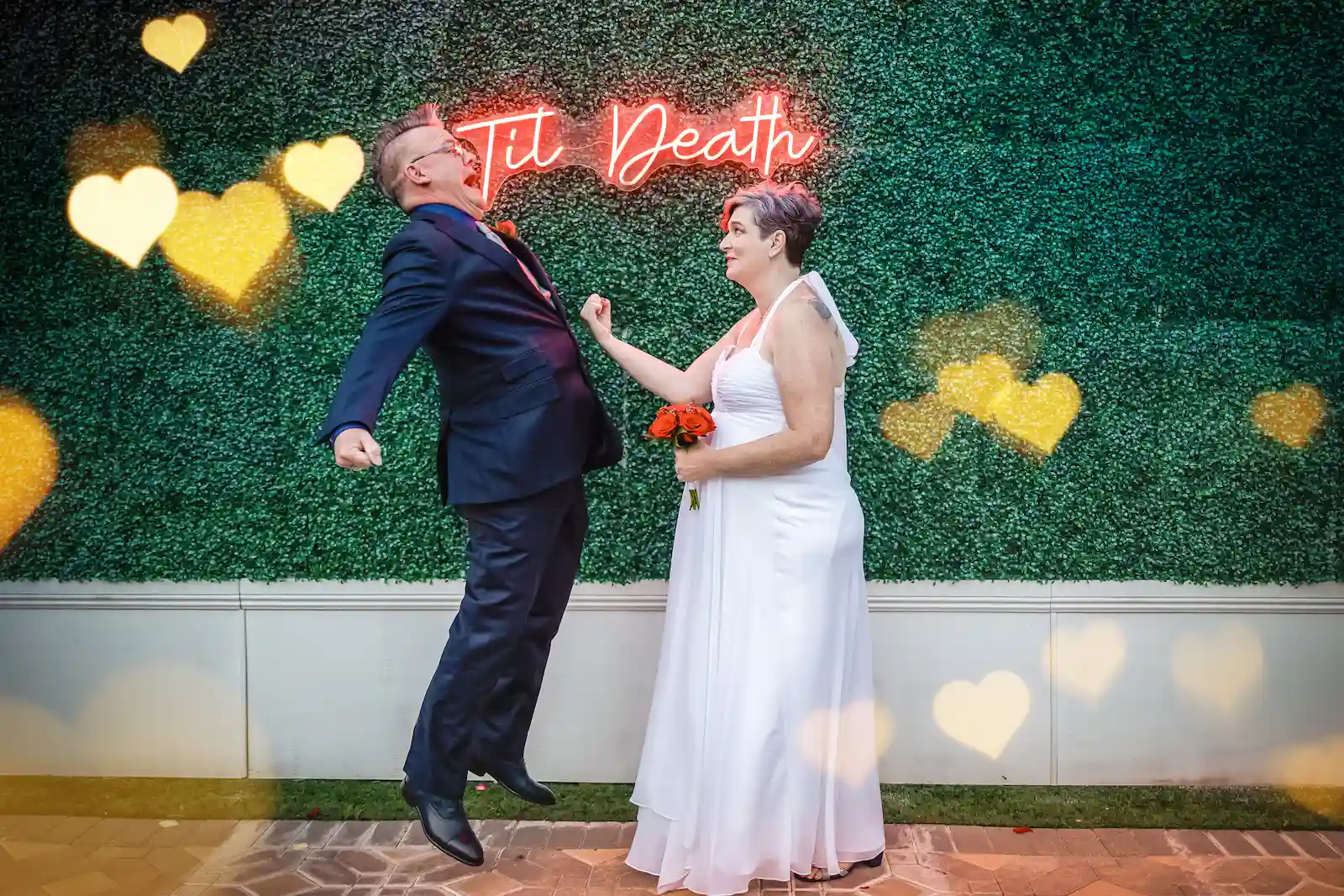 Playful newlyweds celebrating in front of the Til Death neon hedge wall at Viva Las Vegas Wedding Chapel.