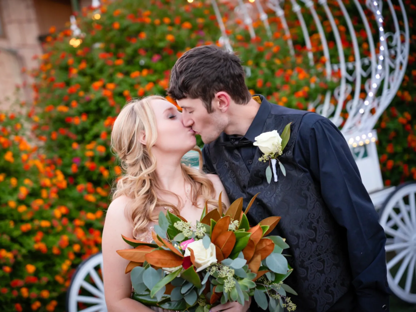 Bride and groom kissing in front of orange flowering bushes and Cinderella carriage at Viva Las Vegas Wedding Chapel.