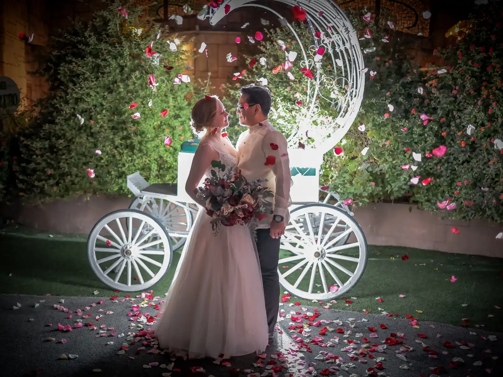 A bride and groom share a romantic moment at night next to a glowing white Cinderella-style carriage, with red and white rose petals scattered on the ground.