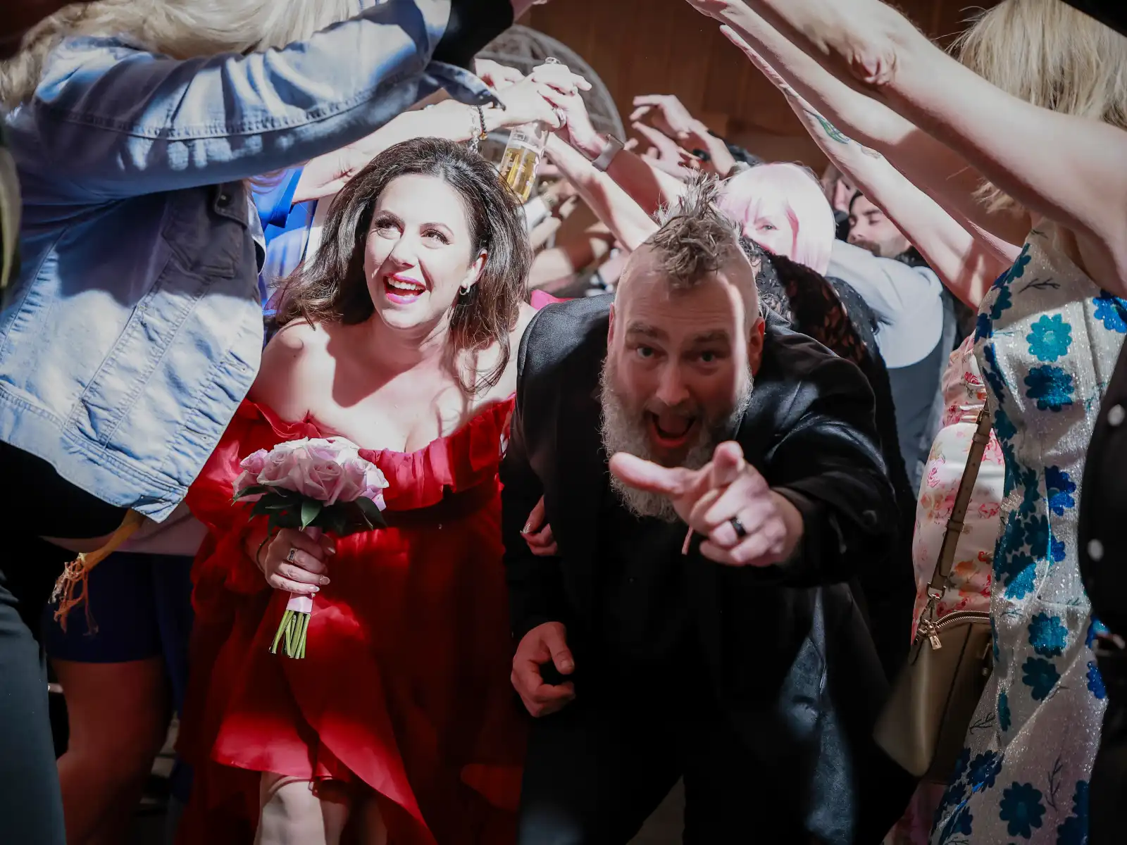 A bride in a striking red dress and a groom with a mohawk joyfully run through a tunnel of cheering guests at their wedding celebration.