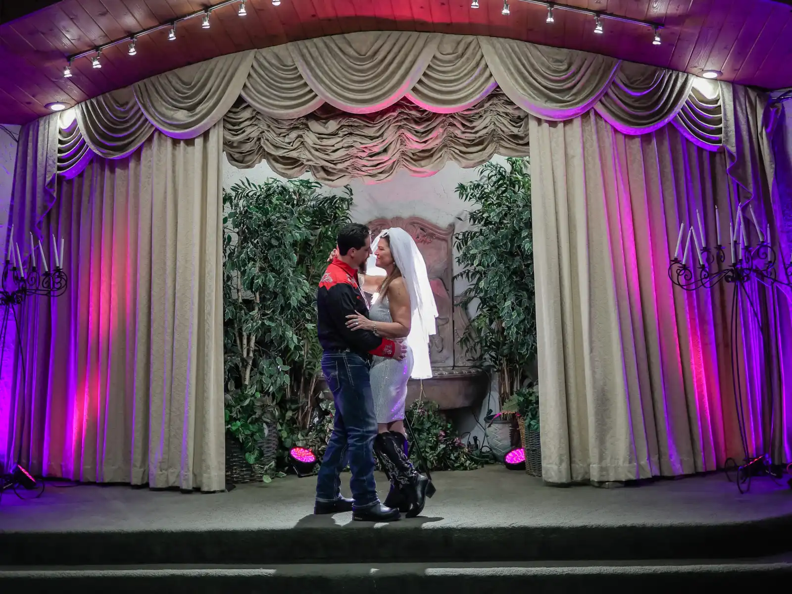 A bride in a white dress and boots embraces her groom, who is wearing a Western-style shirt, on a chapel stage decorated with elegant drapes and purple lighting.