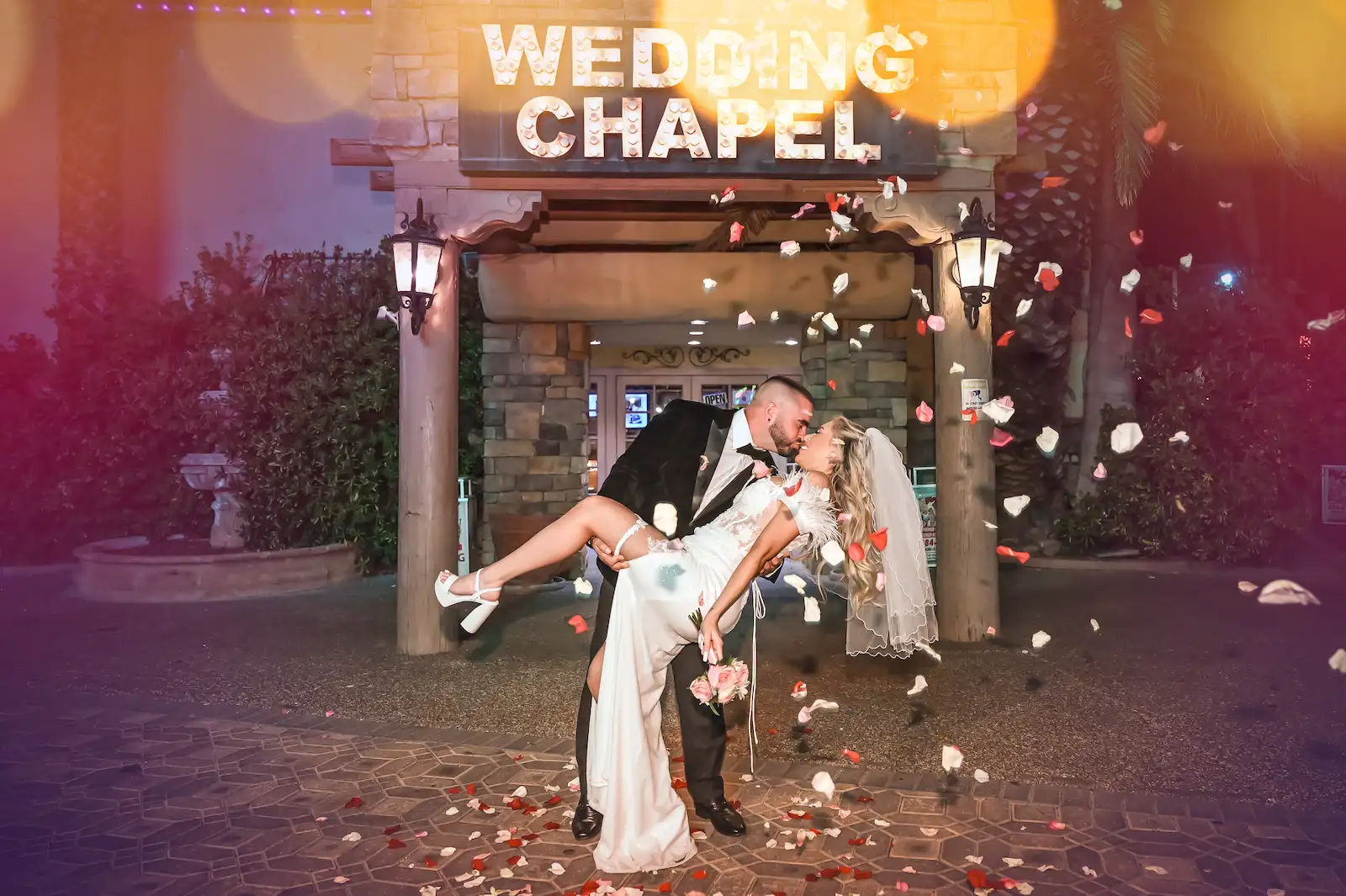 Groom dipping bride for a kiss outside Viva Las Vegas Wedding Chapel entrance with rose petals falling at night.