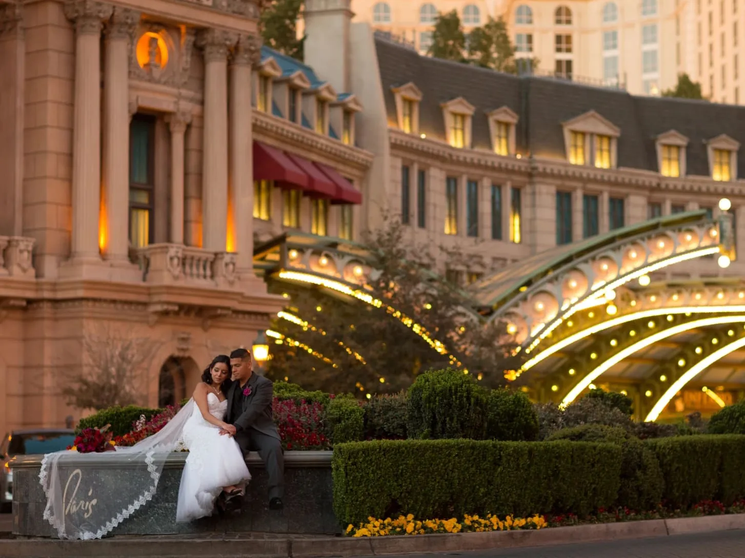 Newlywed couple sitting together in front of Paris Las Vegas hotel entrance at sunset in garden.