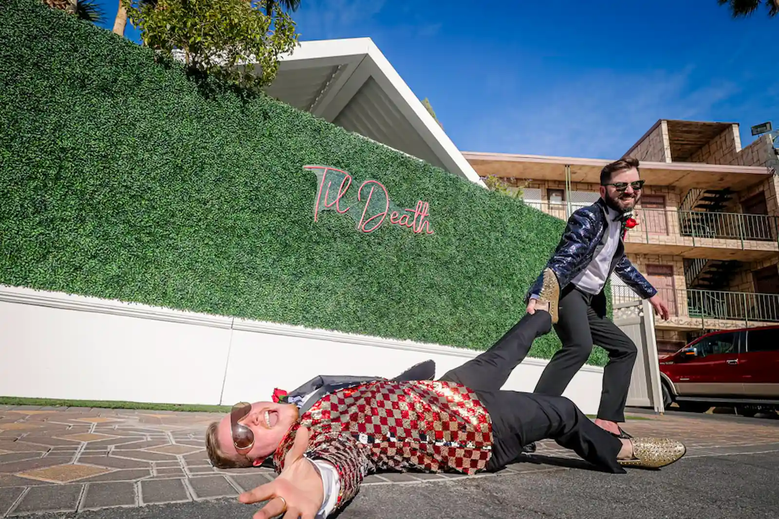 Two grooms in sequin blazers playfully posing in front of the Til Death hedge wall at Viva Las Vegas Wedding Chapel.