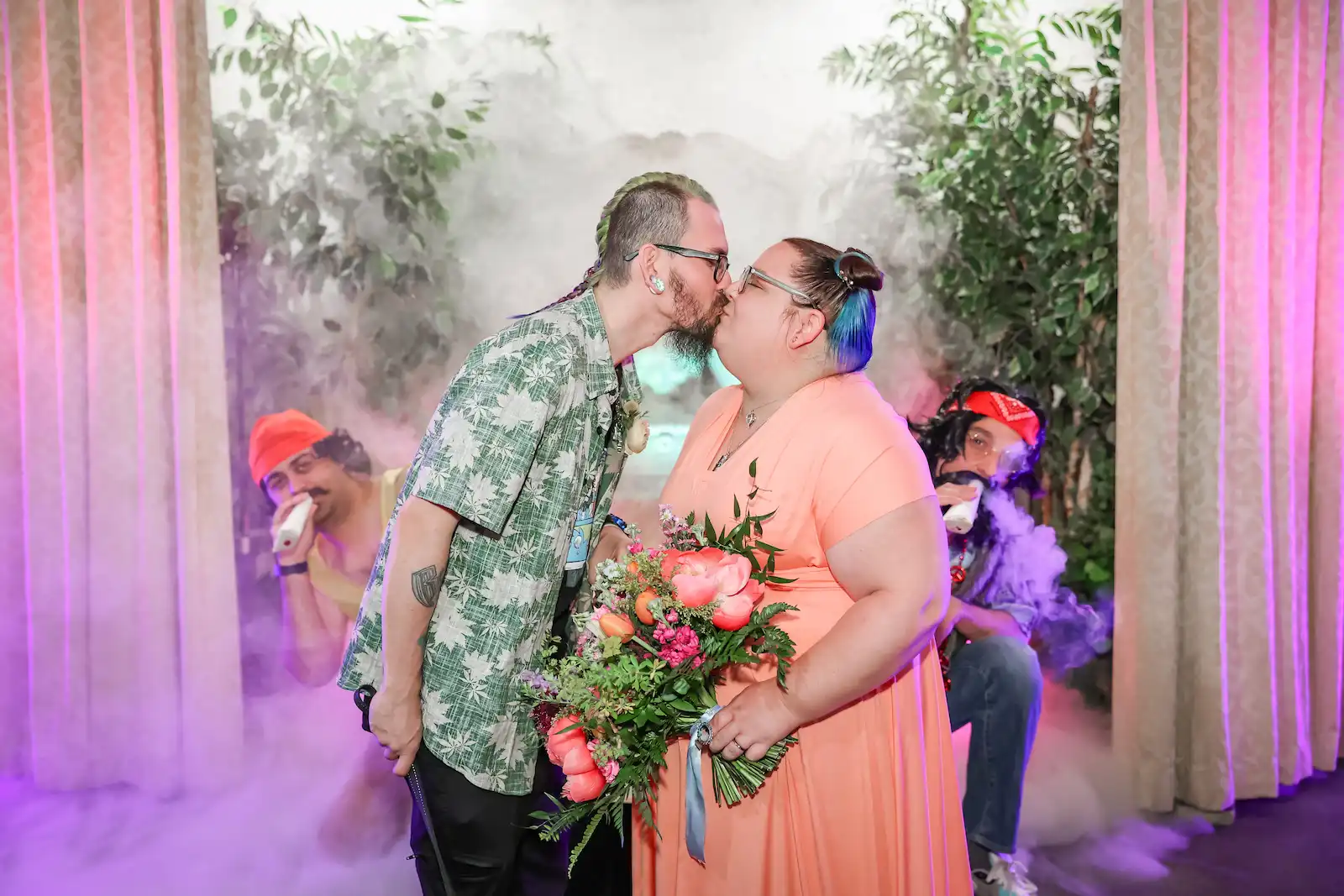 Couple kissing with pink bouquet as Cheech and Chong impersonators pose in background with stage fog at Las Vegas themed wedding
