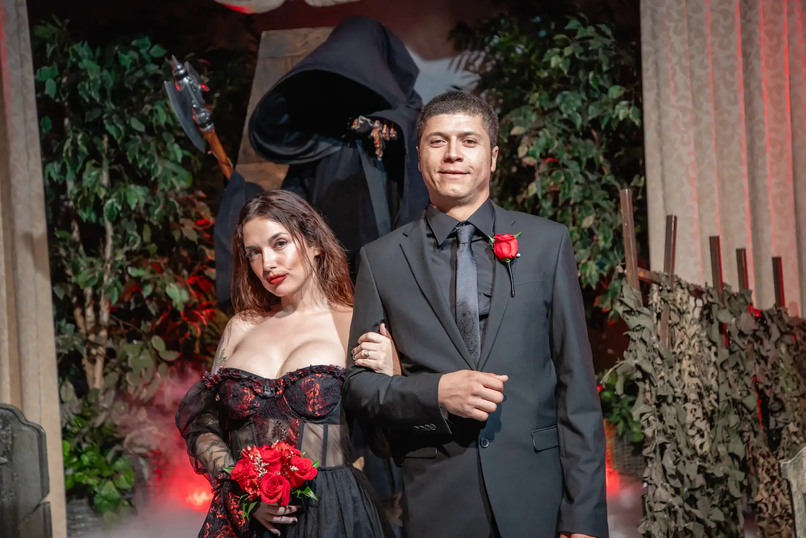 Couple in black and red attire posing with Grim Reaper officiant at a gothic horror-themed Las Vegas wedding chapel with red roses and fog